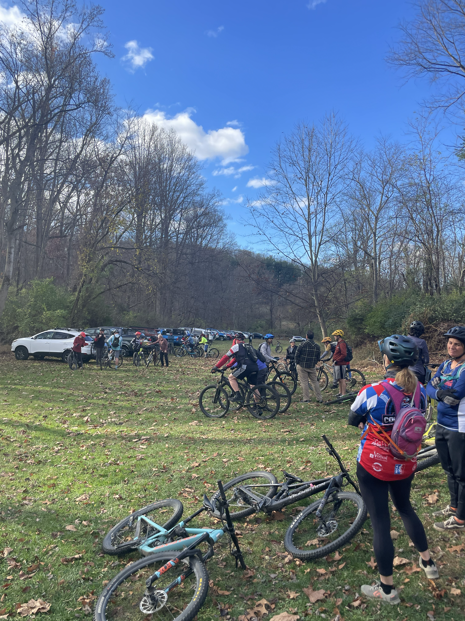 A group of people with bicycles gathering outdoors on a grassy area with fallen leaves, surrounded by tall, leafless trees, under a partly cloudy blue sky.