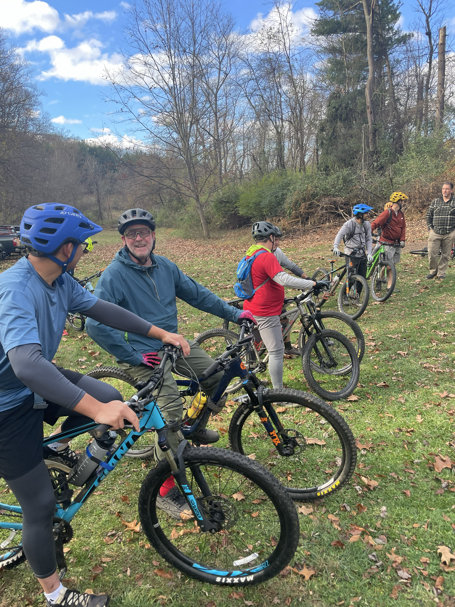 Group of six mountain bikers in helmets, gathered in a park with fallen leaves, bare trees, and a blue sky, preparing or taking a break.