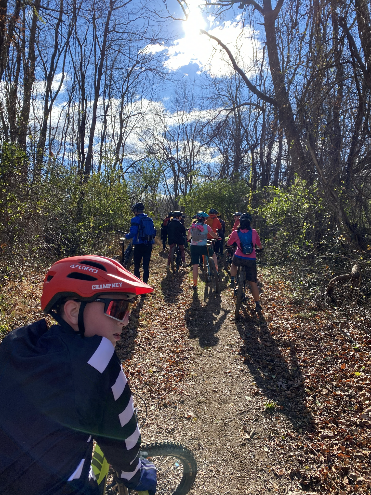 A group of children and adults with bikes and helmets on a dirt trail in a wooded area, with sunlight shining through leafless trees and a partly cloudy blue sky.