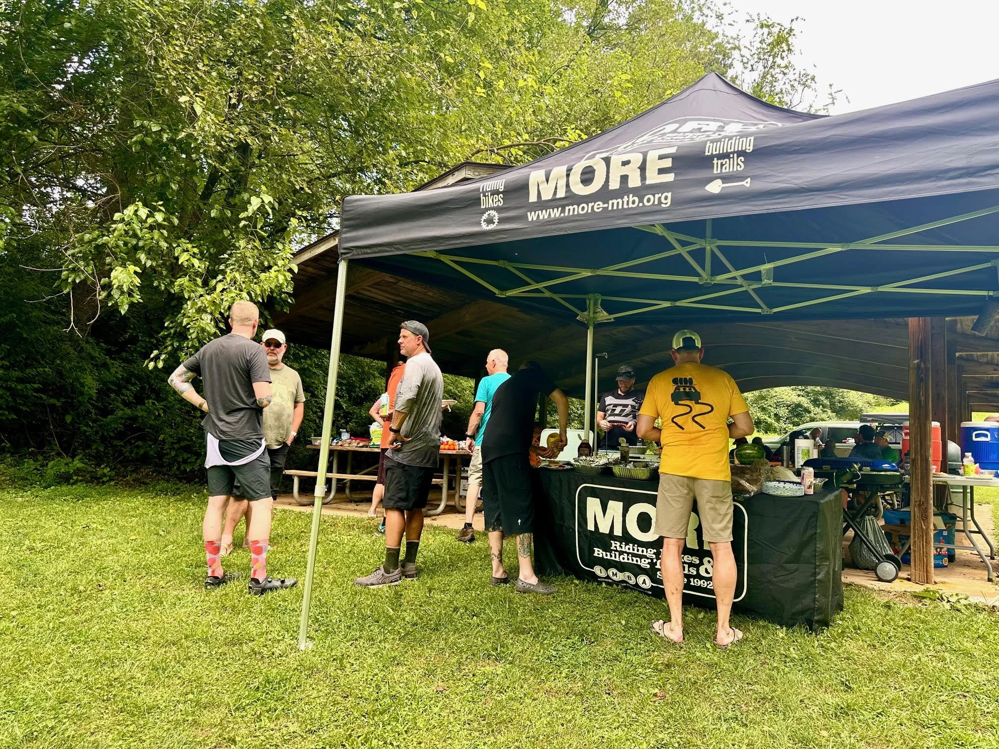 A group of people gathered near a black tent with the logo and website for MORE MTB. The tent is set up outdoors under trees, and there are tables with food and drinks.
