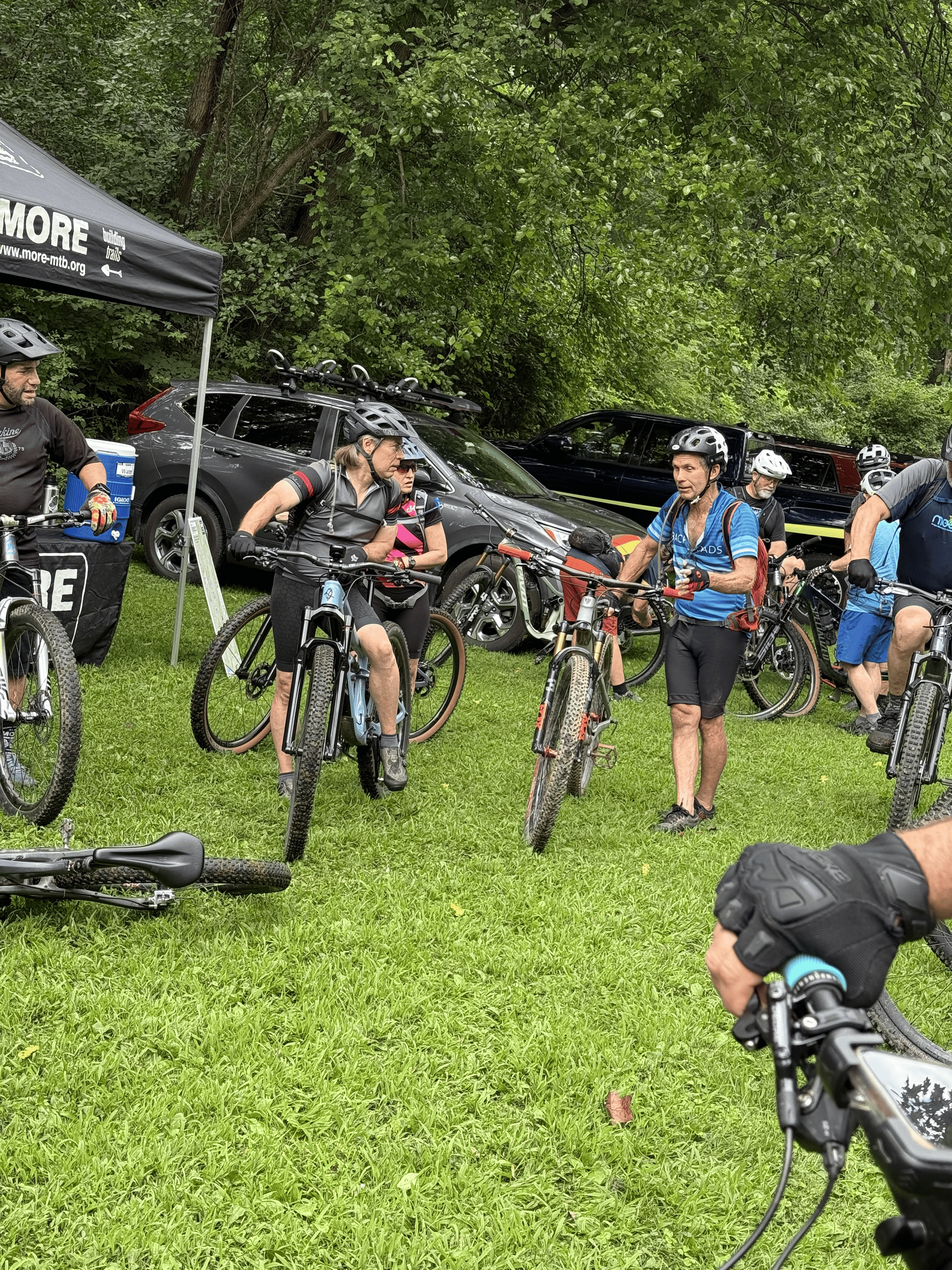 Group of people with bicycles gathered outdoors near a black car and a tent, surrounded by trees and green grass.