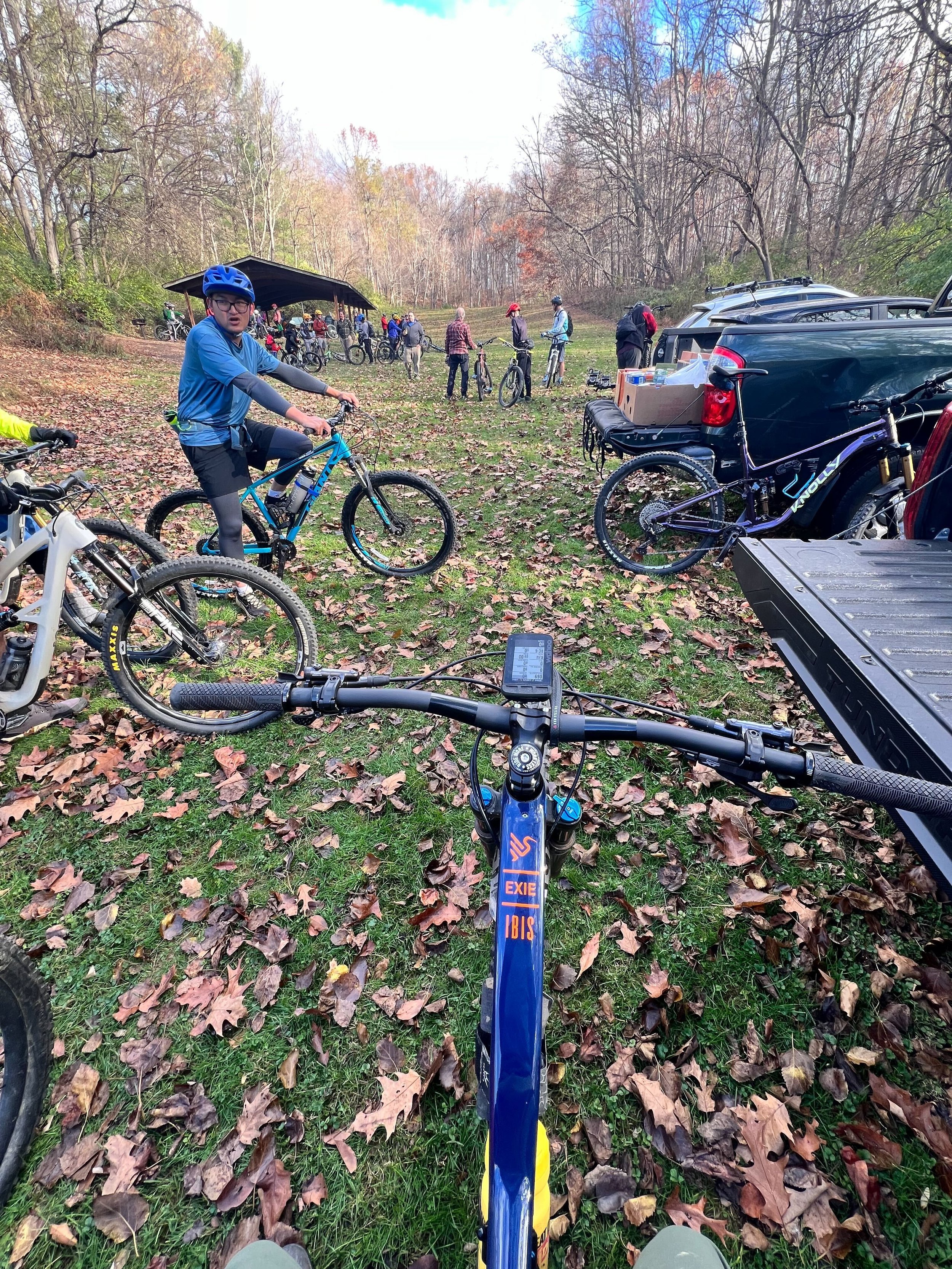 View from a mountain bike on a trail covered with fallen leaves, with several cyclists and vehicles parked along the side, and a wooded area with autumn trees in the background.