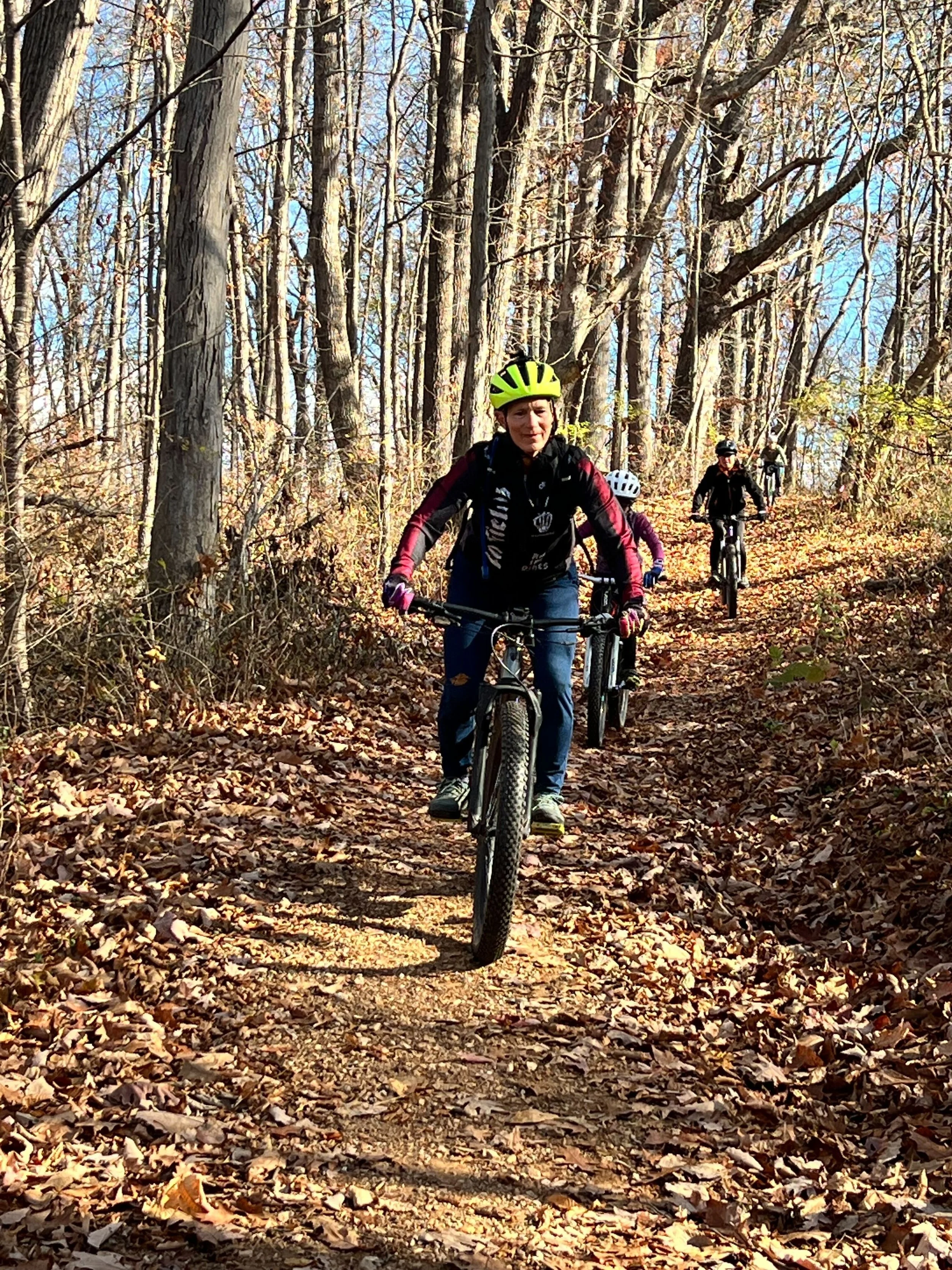 A group of people mountain biking on a leaf-covered forest trail in autumn, with trees on either side and a clear sky.