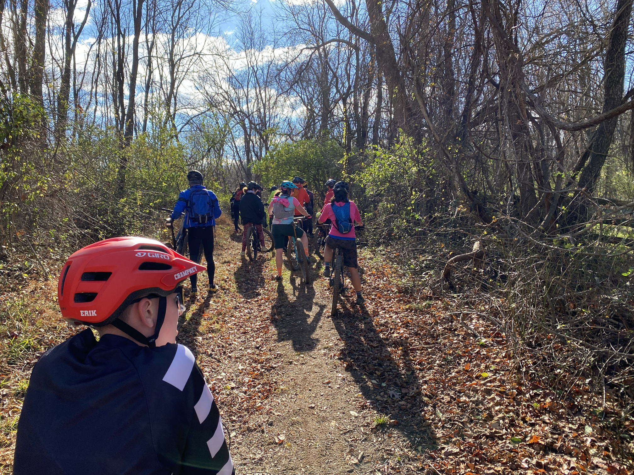 Children wearing helmets and backpacks riding bicycles on a forest trail, surrounded by trees and fallen leaves, under partly cloudy sky.
