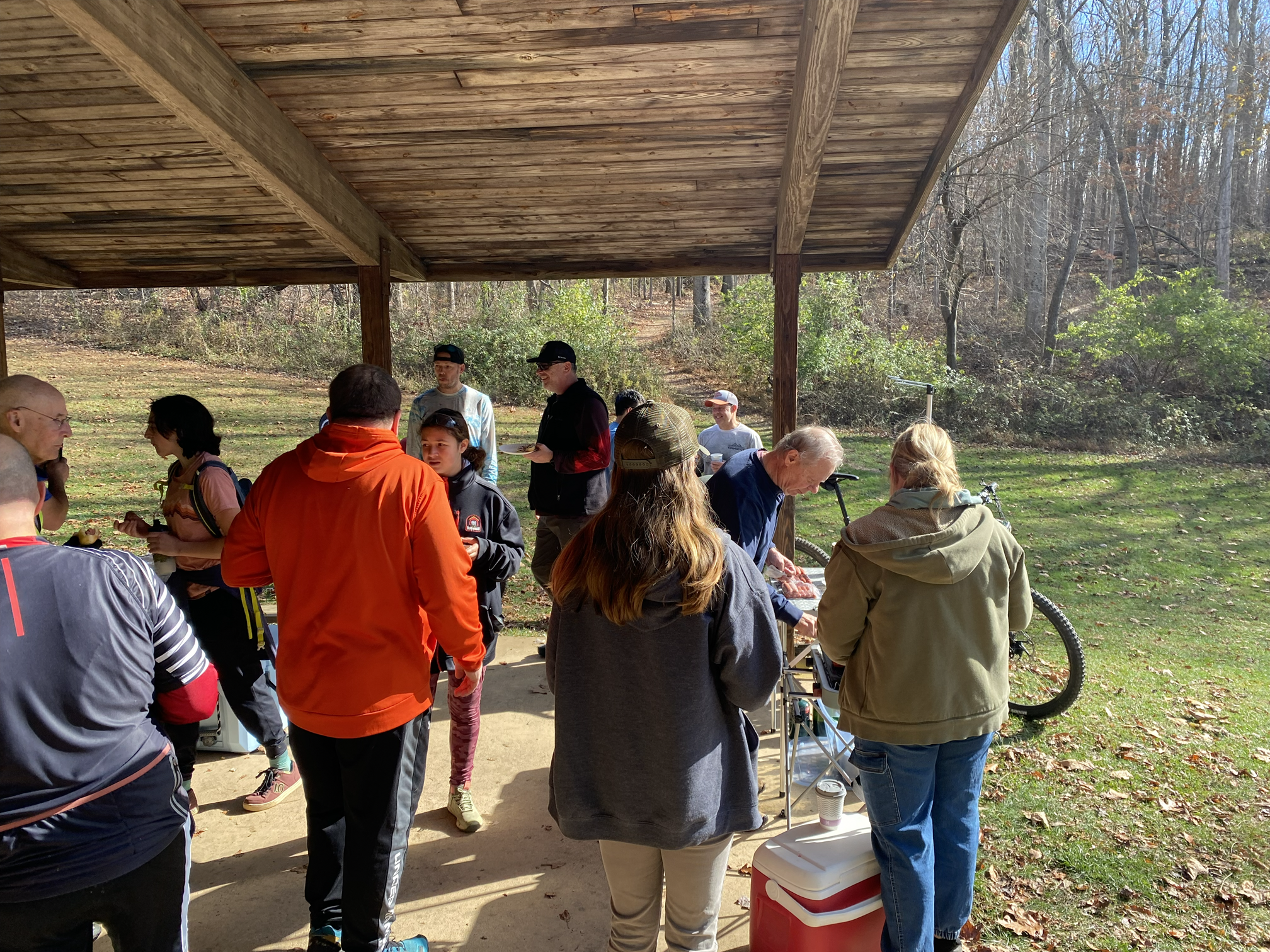 People gathered under a wooden pavilion outdoors, possibly at a park, with a wooded background. Some people are standing, others are sitting or engaging in activities around tables, with a person serving food or drinks.
