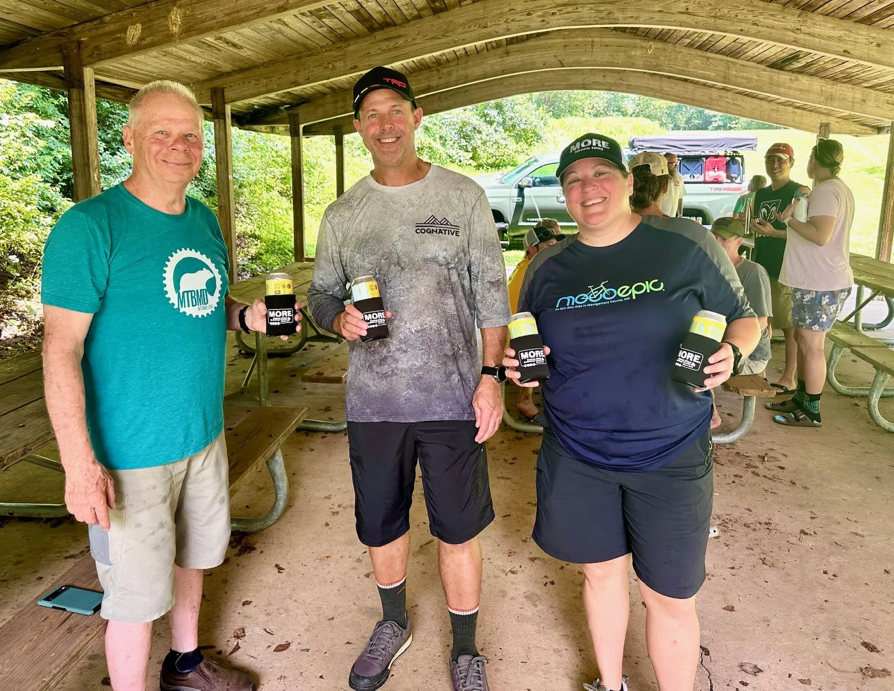 Three people standing under a wooden shelter, smiling and holding drinks, with other people in the background at an outdoor event.