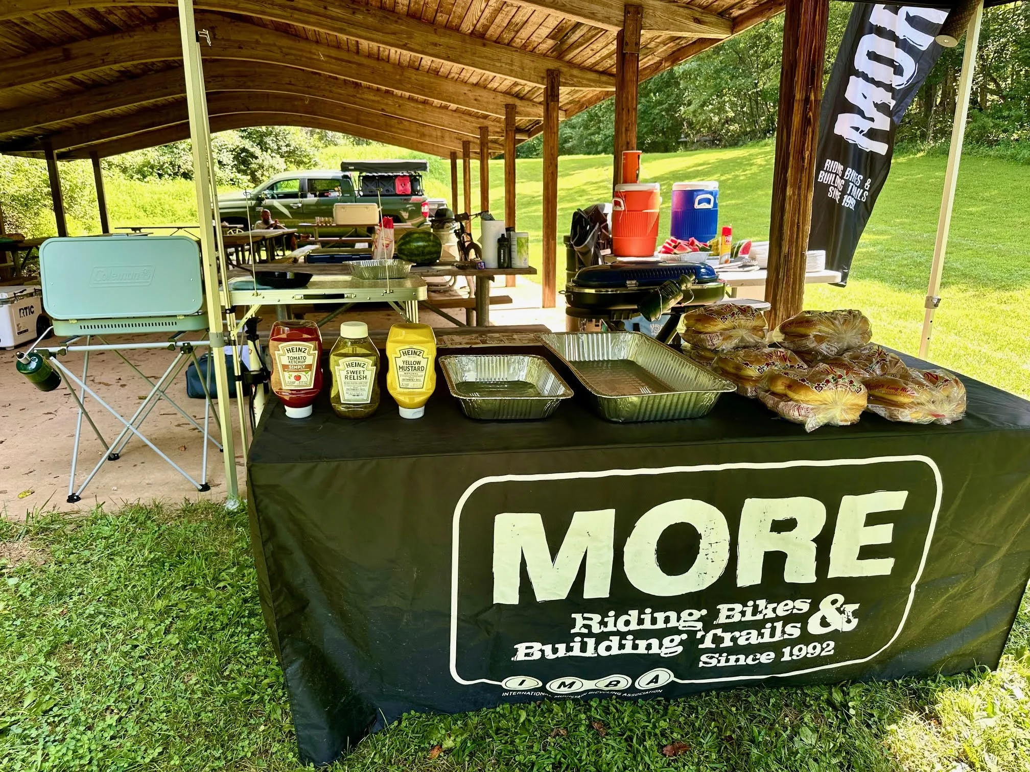 A covered outdoor area with picnic tables set up for a gathering or barbecue. There are condiments, drinks, and baked goods on a table in the foreground. Cars are parked behind the area, and a flag with black background and white text advertising rid