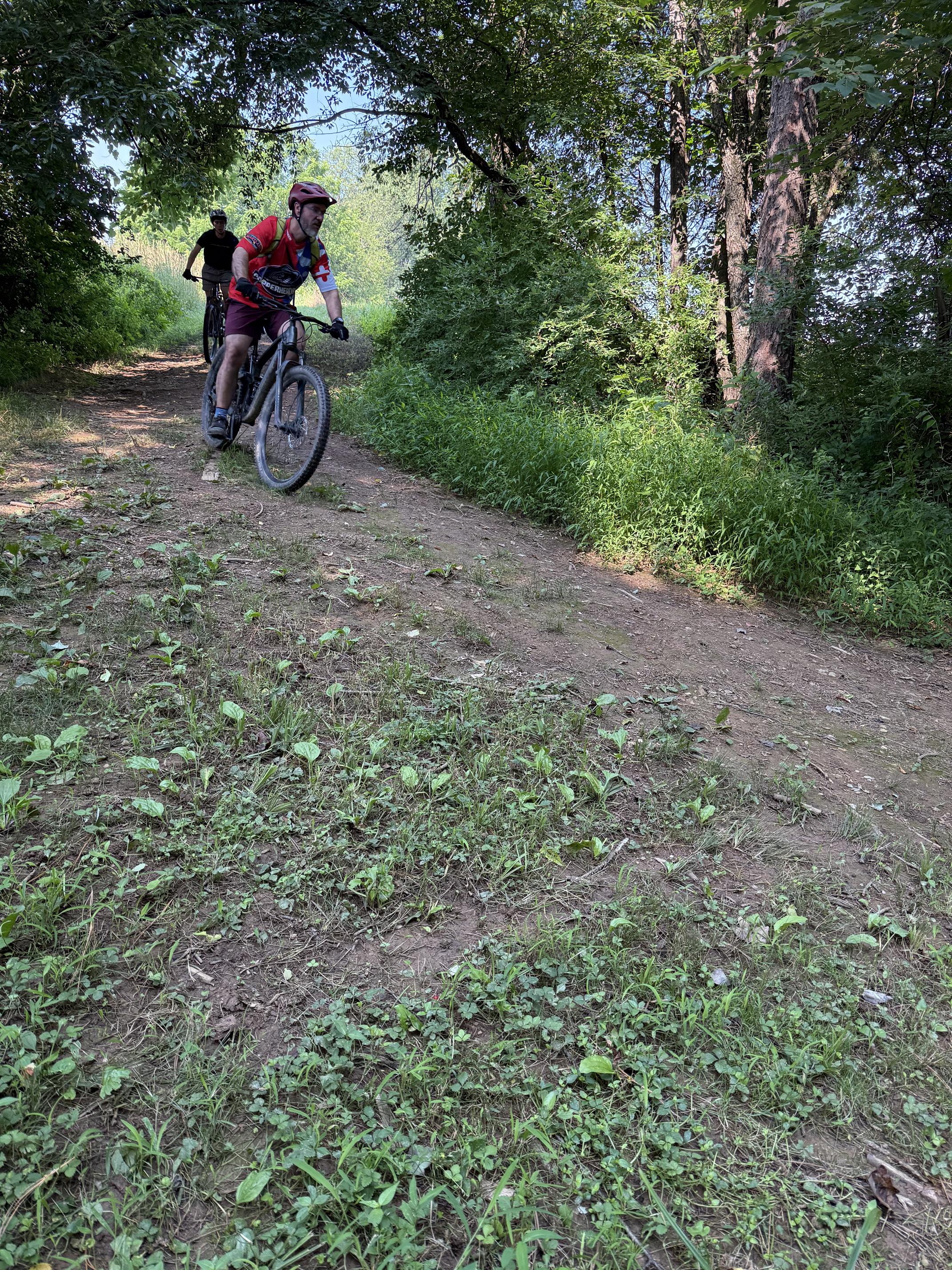 Two people riding bicycles on a dirt trail through a green, wooded area with tall trees and bushes.