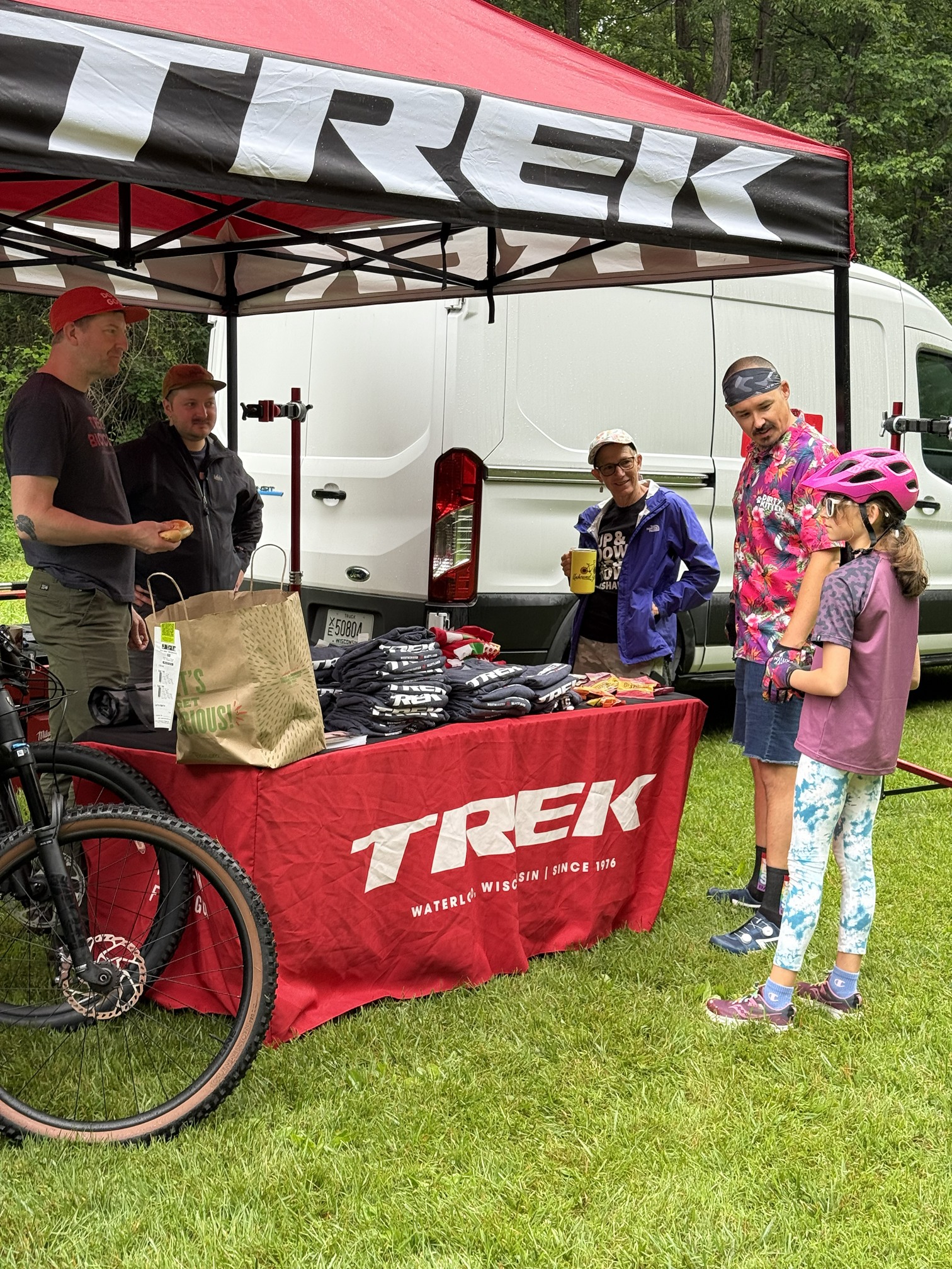People gathered around a merchandise table at a Trek event, outdoors on a grassy area near trees, with parked vehicles in the background. The table is covered with Trek apparel, and a red canopy with the Trek logo provides shade.
