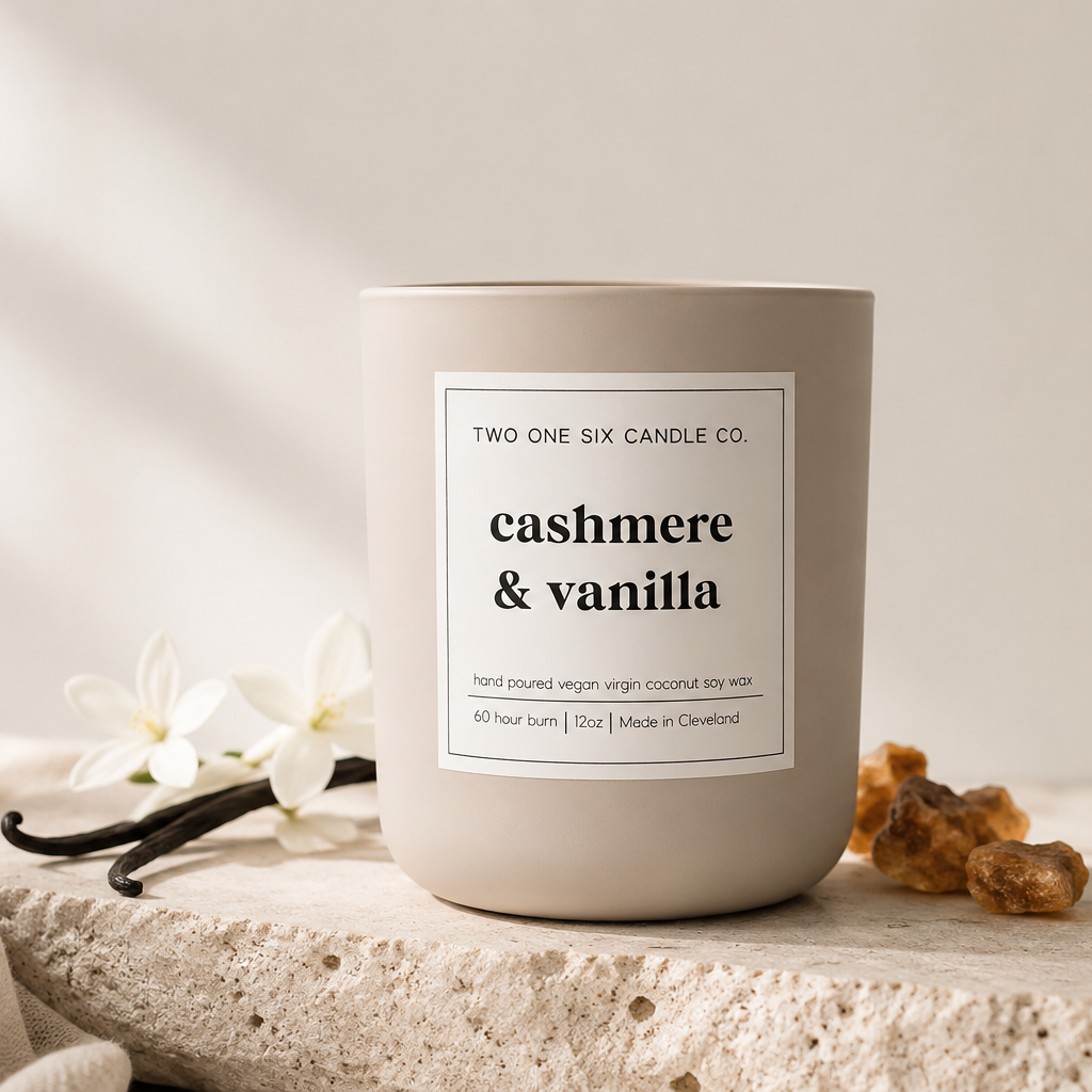 A beige candle in a gray container labeled 'cashmere & vanilla' from Two One Six Candle Co. on a textured surface with vanilla pods, white flowers, and amber stones beside it, with soft natural light in the background.