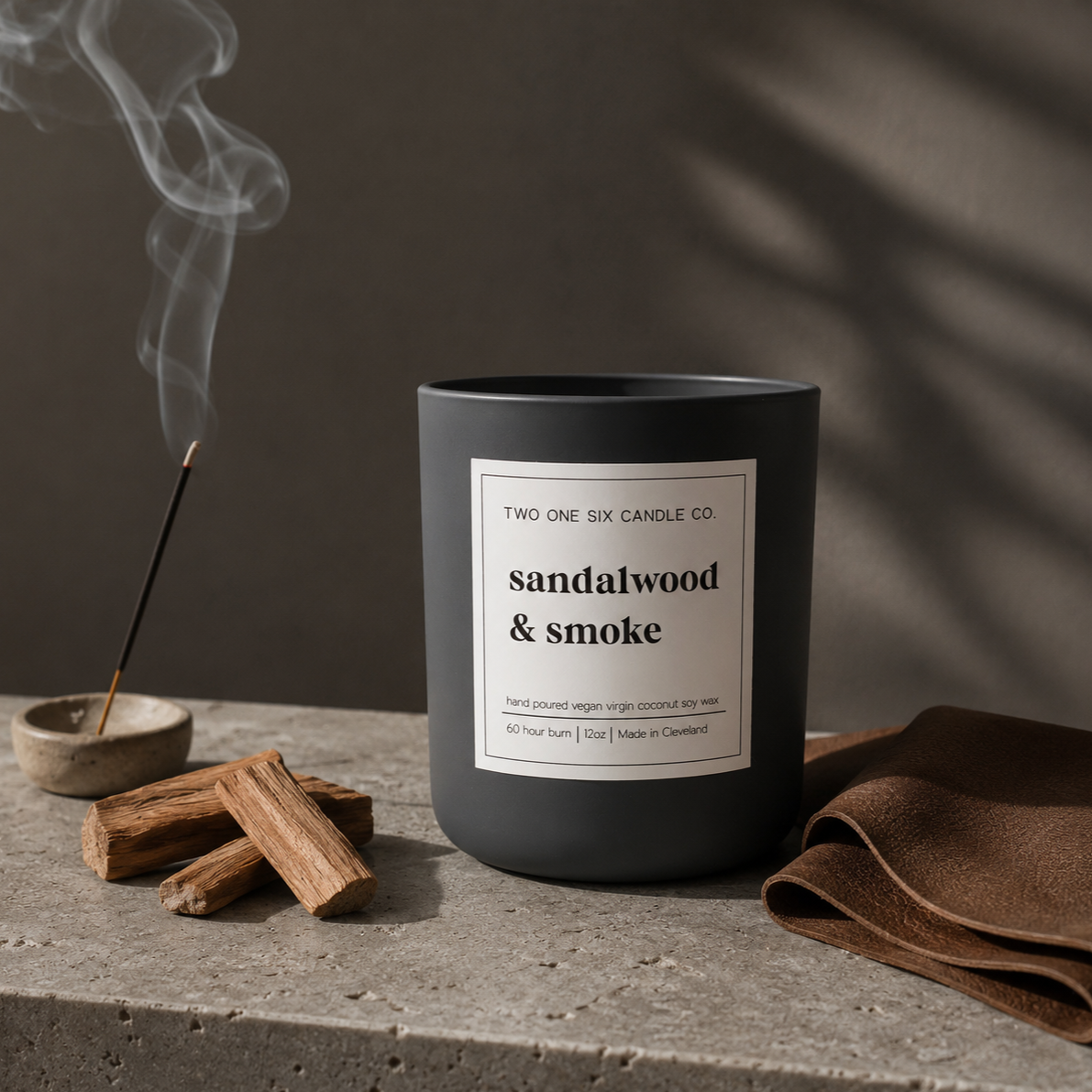 A black candle labeled "sandalwood & smoke" from Two One Six Candle Co. on a concrete surface, with a burning incense stick, pieces of sandalwood, and a folded brown leather piece nearby. Smoke rises from the incense.