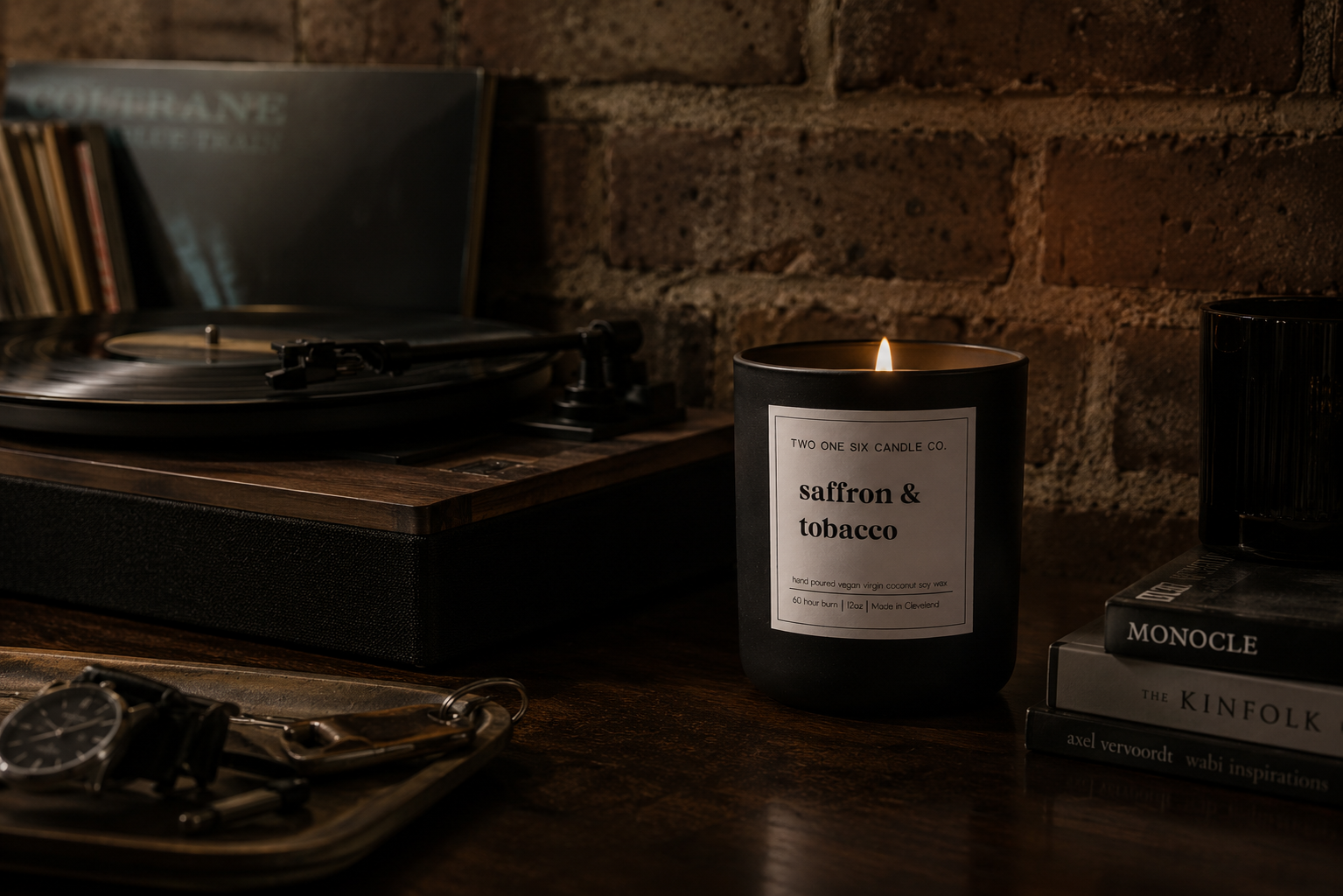 A black candle labeled 'saffron & tobacco' lit beside a record player and a stack of books on a wooden surface against a brick wall.