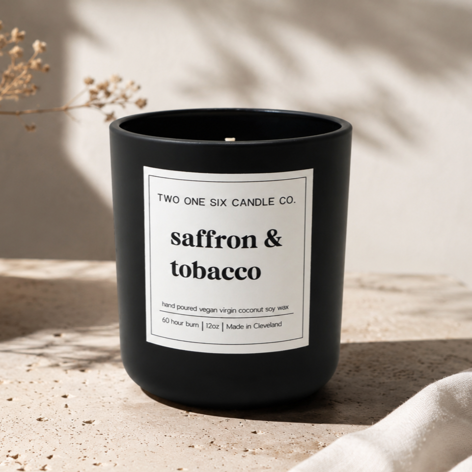 Black candle jar labeled 'saffron & tobacco' by Two One Six Candle Co., resting on a textured beige surface with dried flowers nearby.