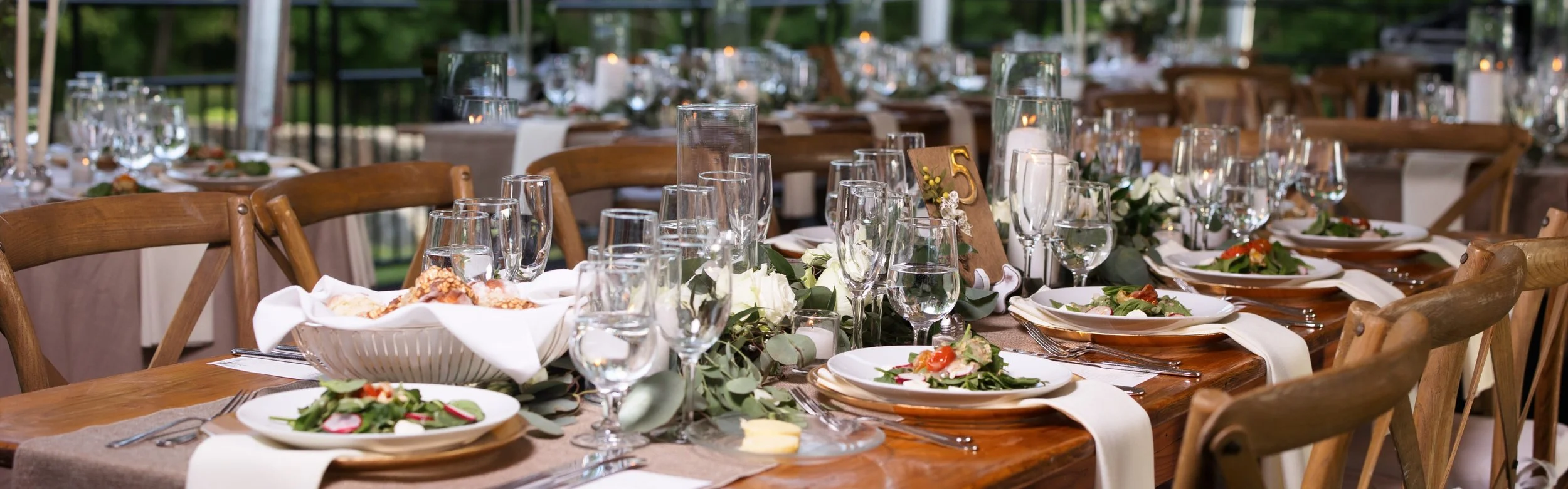 Elegant outdoor banquet table set with white plates, silverware, multiple wine glasses, floral centerpieces, and wooden chairs, prepared for a formal event.
