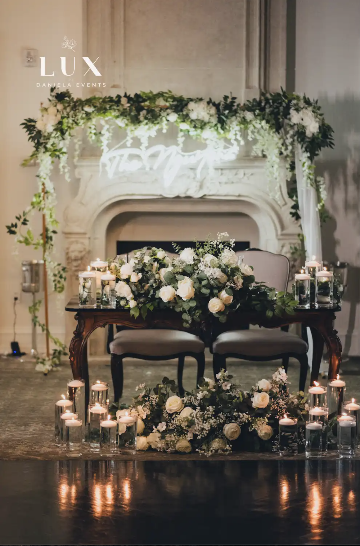 Elegant wedding setup with a fireplace decorated with white flowers and greenery, a wooden table with floral arrangements, candles in glass holders, and a lit neon sign