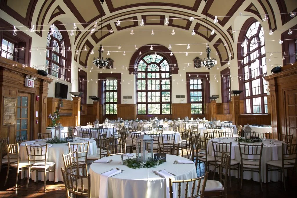 Elegant banquet hall decorated with round tables covered in white tablecloths, surrounded by gold chairs, and adorned with green foliage centerpieces. String lights hang from the ceiling with chandeliers, and large windows let in natural light. Wooden paneling decorates the walls.
