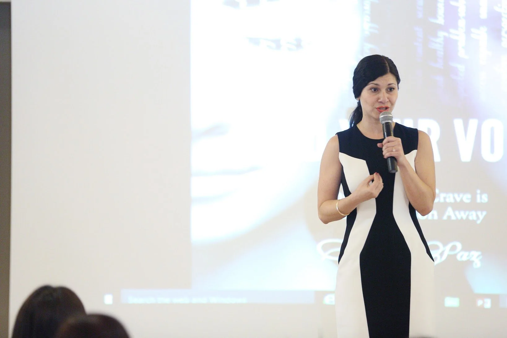 Woman in black and white dress speaking into a microphone at a presentation with a projector screen in the background.