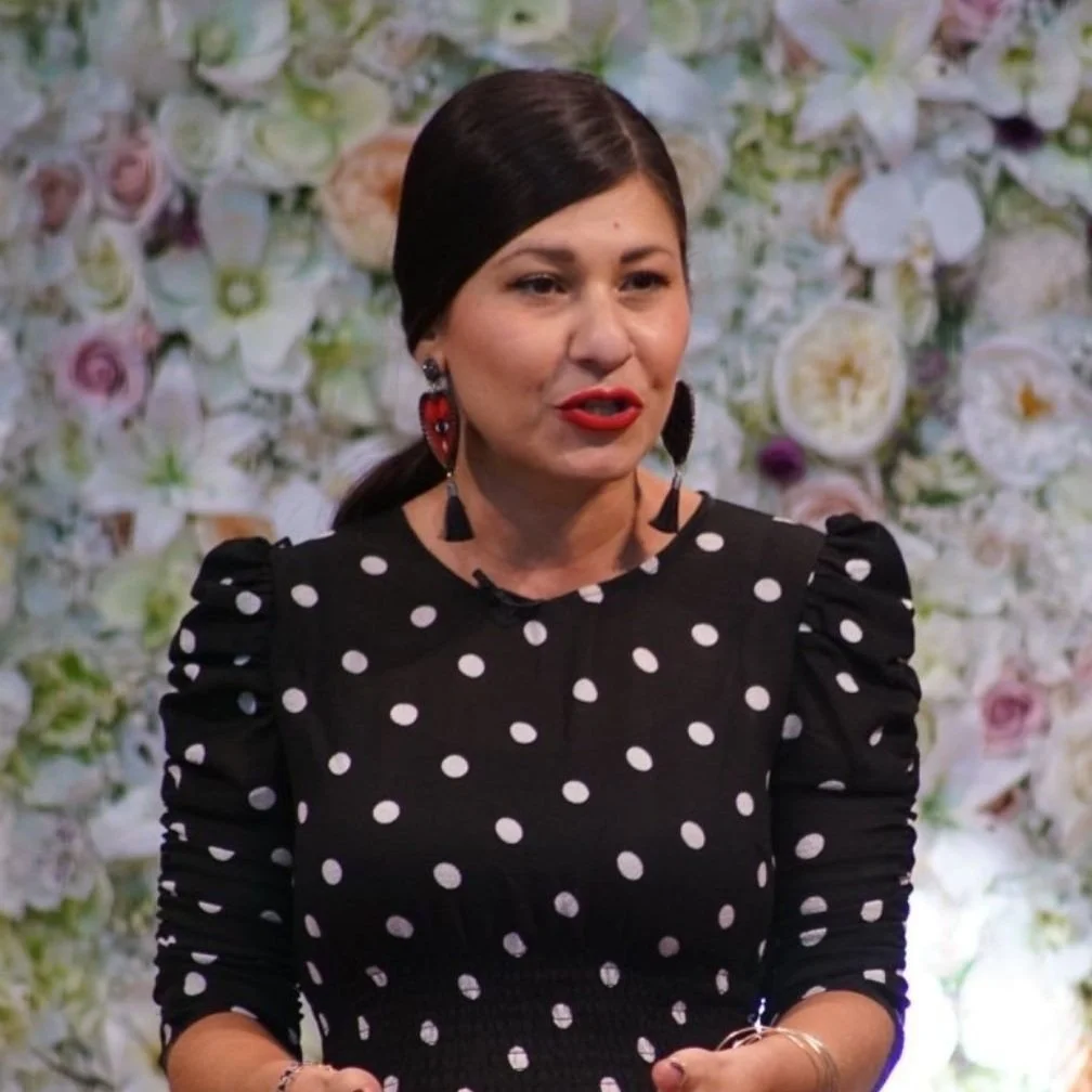 Woman in black polka dot dress speaking against a floral background.