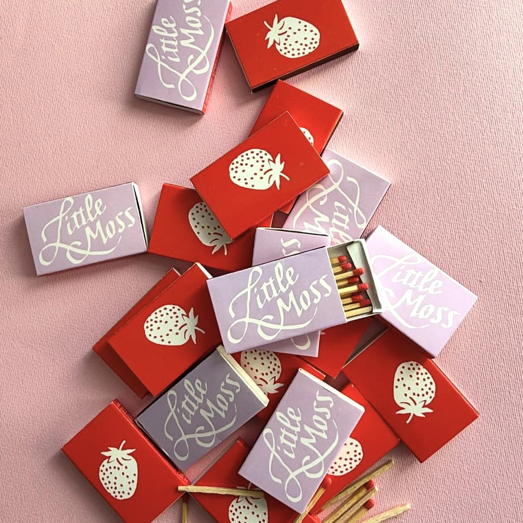 An assortment of pink and lavender matchbooks that say "Little Moss" on one side and have a bold graphic of a strawberry on the other.