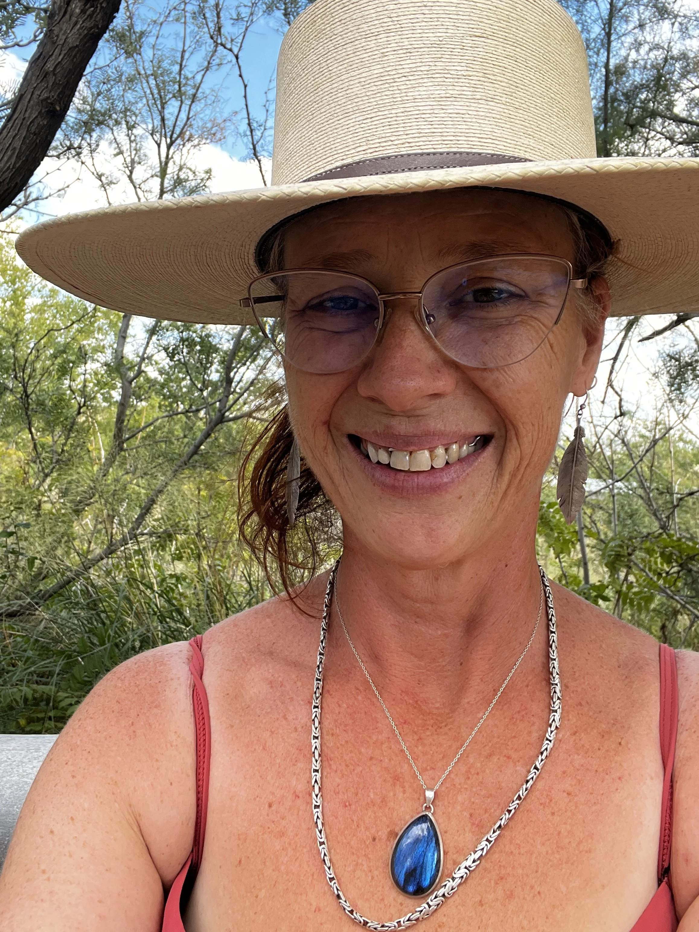 A woman smiling outdoors, wearing a wide-brimmed straw hat, glasses, feather earrings, a necklace with a blue pendant, and a coral-colored top.