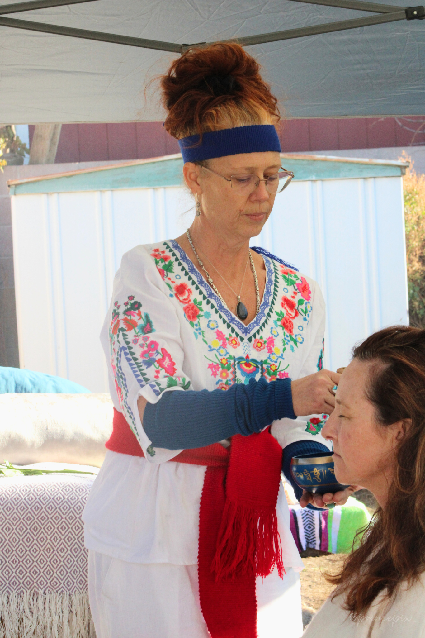 A woman wearing a traditional embroidered blouse, a blue headband, glasses, and jewelry, is performing a ceremonial act with a small bowl held over the forehead of another woman with long brown hair. They are outdoors under a tent with blankets and a shed in the background.