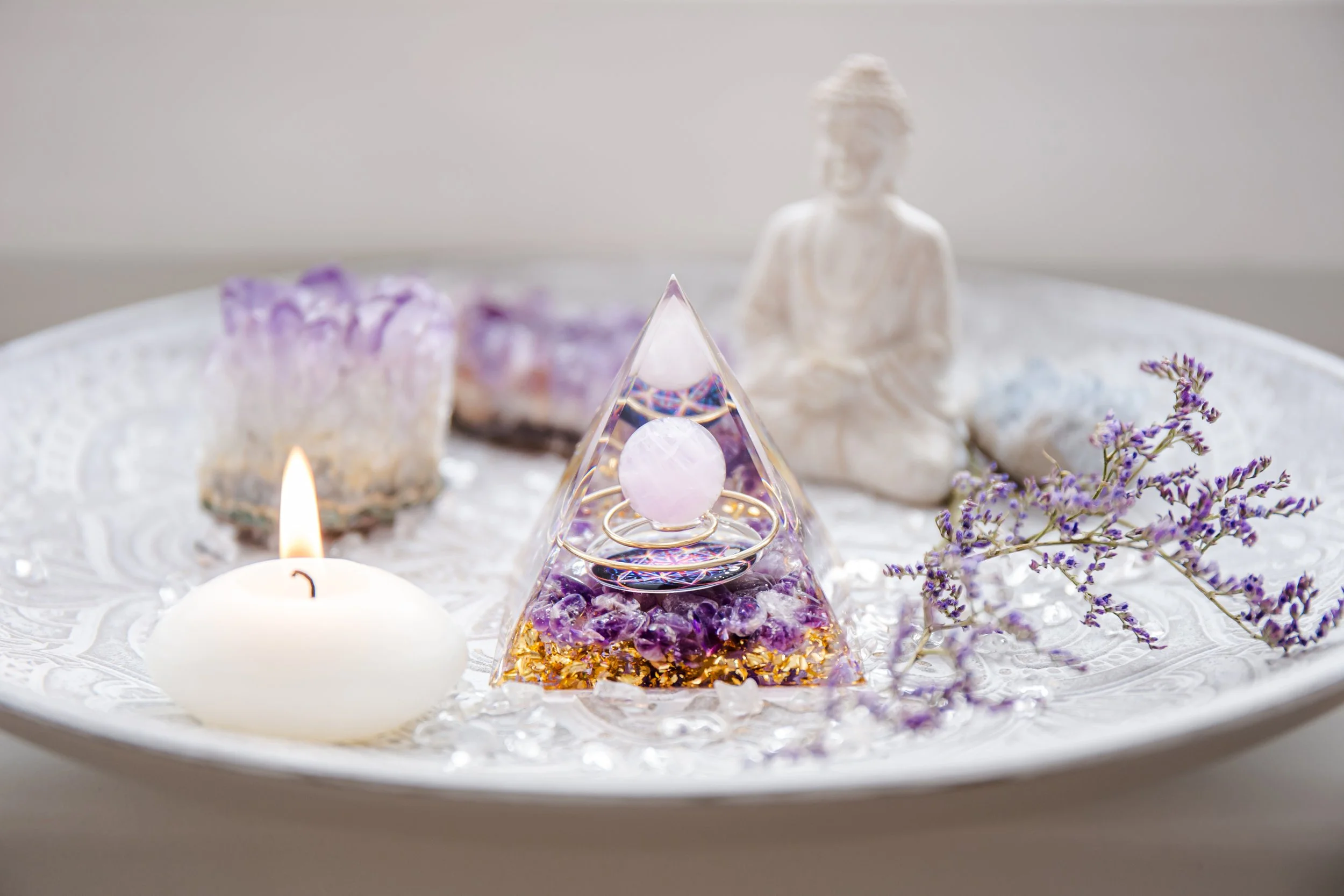 A decorative display on a white surface, featuring a lit white candle, a purple and gold gem-like ornament with jewelry inside, a small white statue of a seated person, purple flowers, and purple mineral stones in the background.