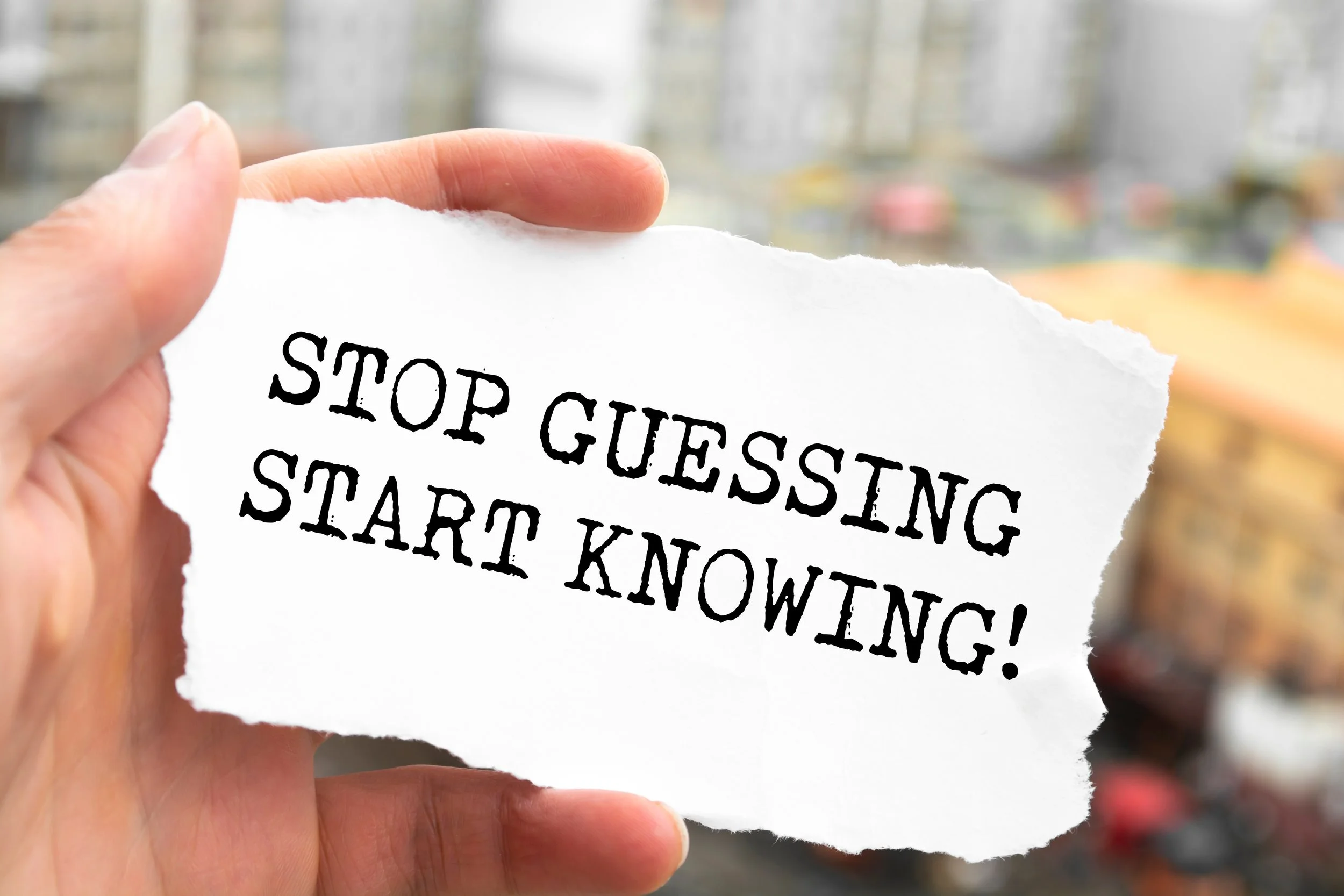 Hand holding a torn piece of white paper with the message 'STOP GUESSTIM START KNOWING!' written in bold black text.