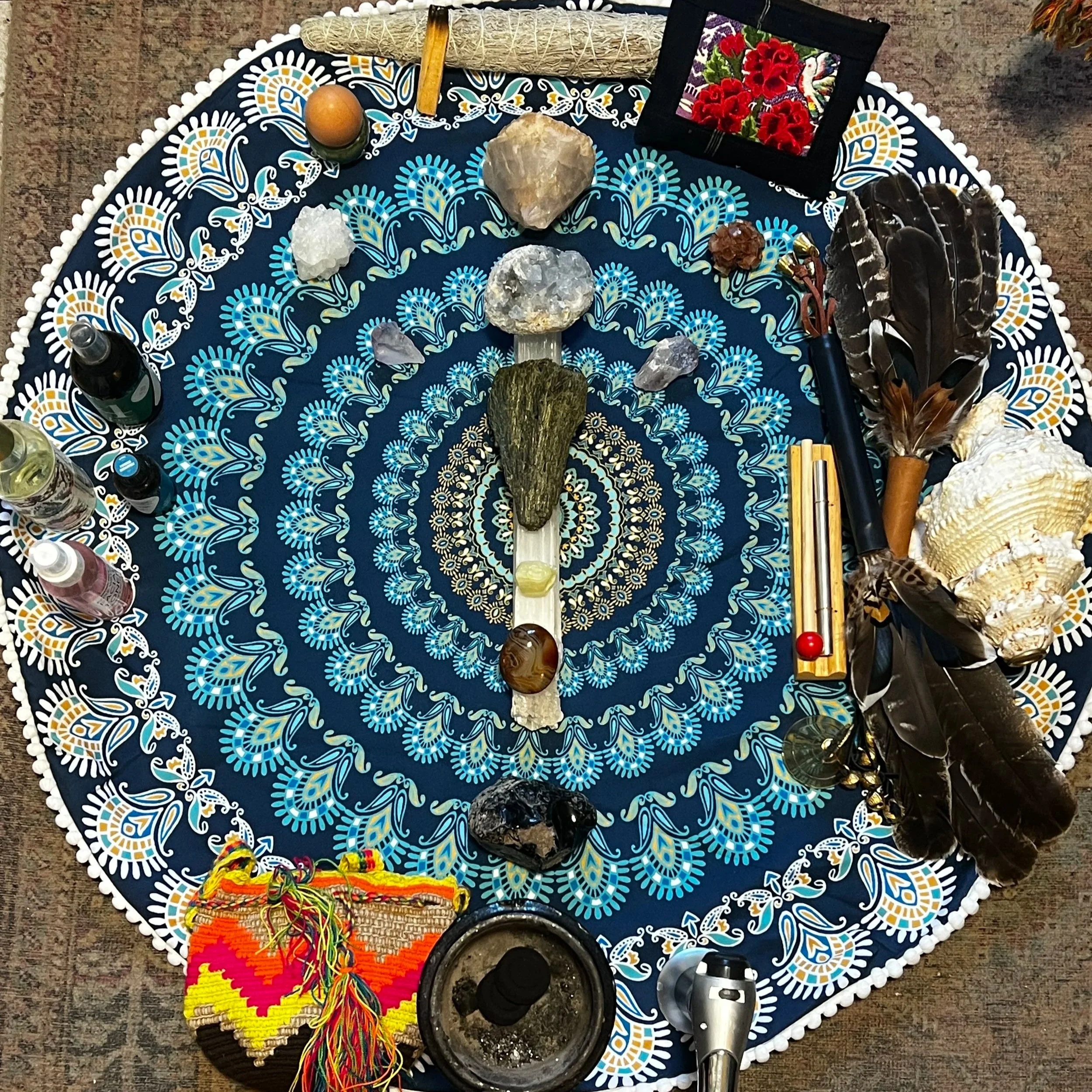 A circular, decorative cloth with blue, white, and yellow patterns on a surface. In the center, there are various stones and crystals placed in a symmetrical arrangement. Around the perimeter, there are bottles, a small framed embroidered picture, feathers, shells, a drum, a guiro instrument, a small colorful woven cloth, and other spiritual or ritual objects.