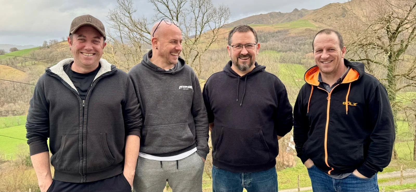 Four men standing outdoors in front of scenic hills, wearing casual hoodies and smiling, with a background of trees and green landscape.