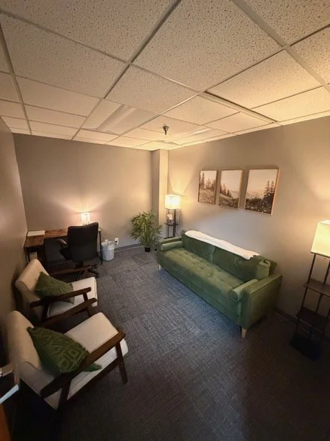 A cozy waiting room with a green sofa, two armchairs, a desk with an office chair, and a small potted plant. Wall art and lamps provide warm lighting.