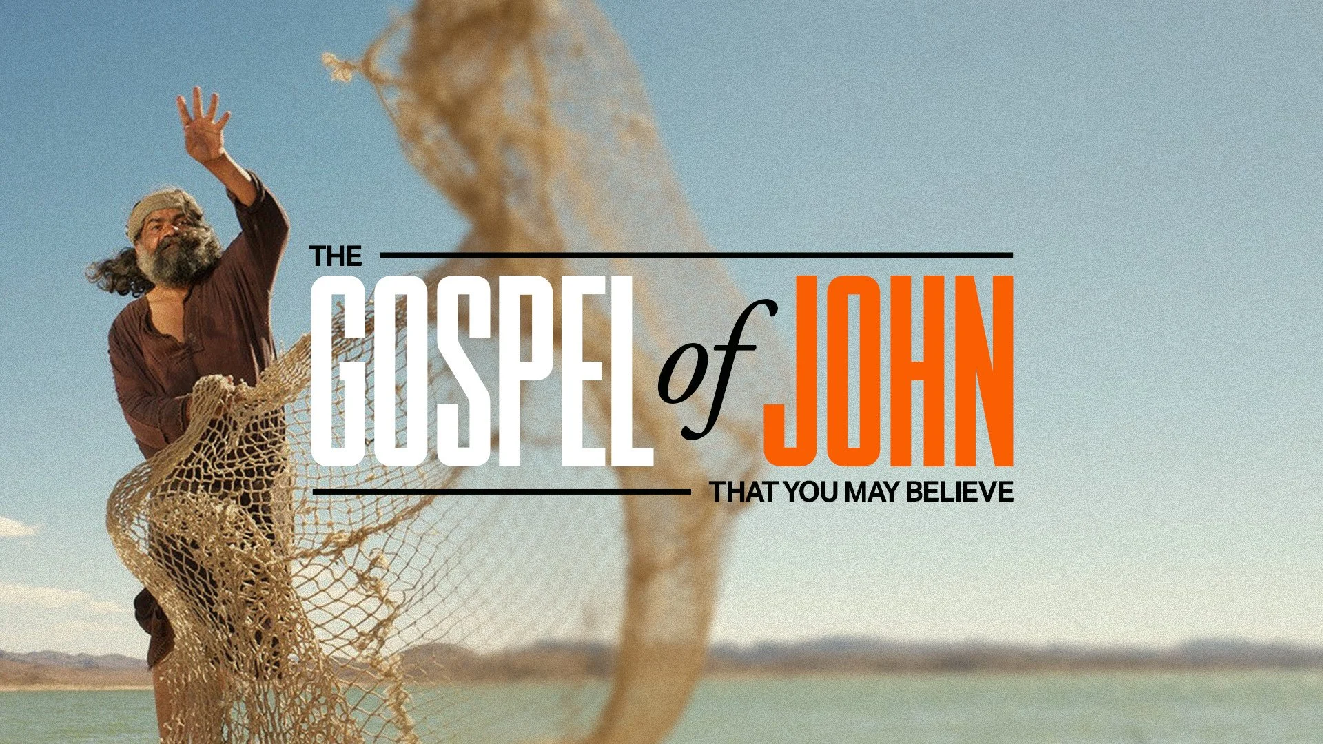 A man with long hair and a beard, wearing a beige cap and brown shirt, standing outdoors by a body of water, holding a fishing net, with mountains in the background and the sky clear.