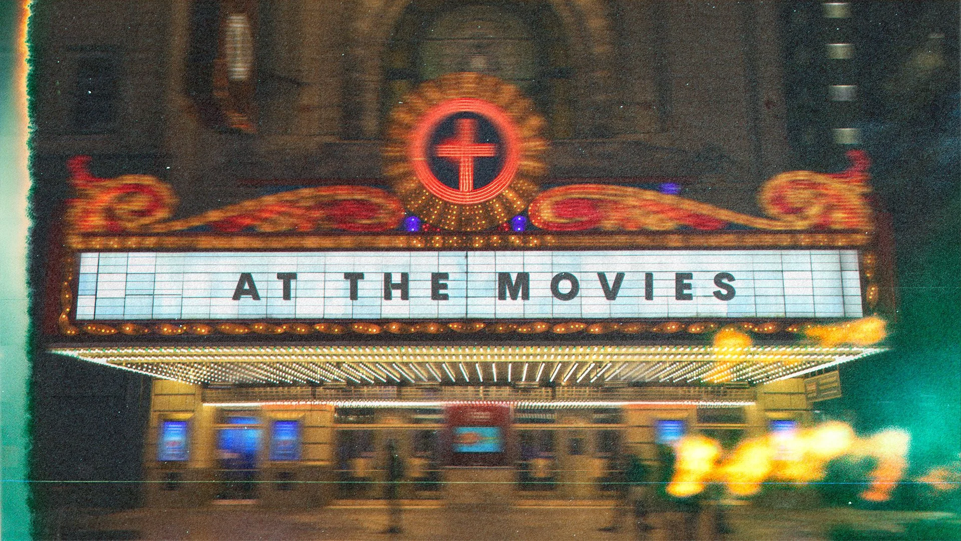 Marquee sign with the words 'AT THE MOVIES' at night outside a theater, with decorative lights and a Christian cross symbol above.