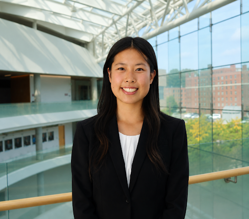 A young woman in a black blazer and white blouse smiling inside a modern building with large glass windows and a view of trees and buildings outside.