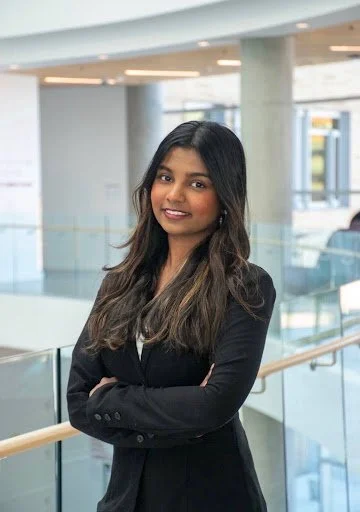 A young woman with long dark hair wearing a black blazer, smiling confidently with her arms crossed, standing in a modern, well-lit office or airport terminal with large windows and glass railings.