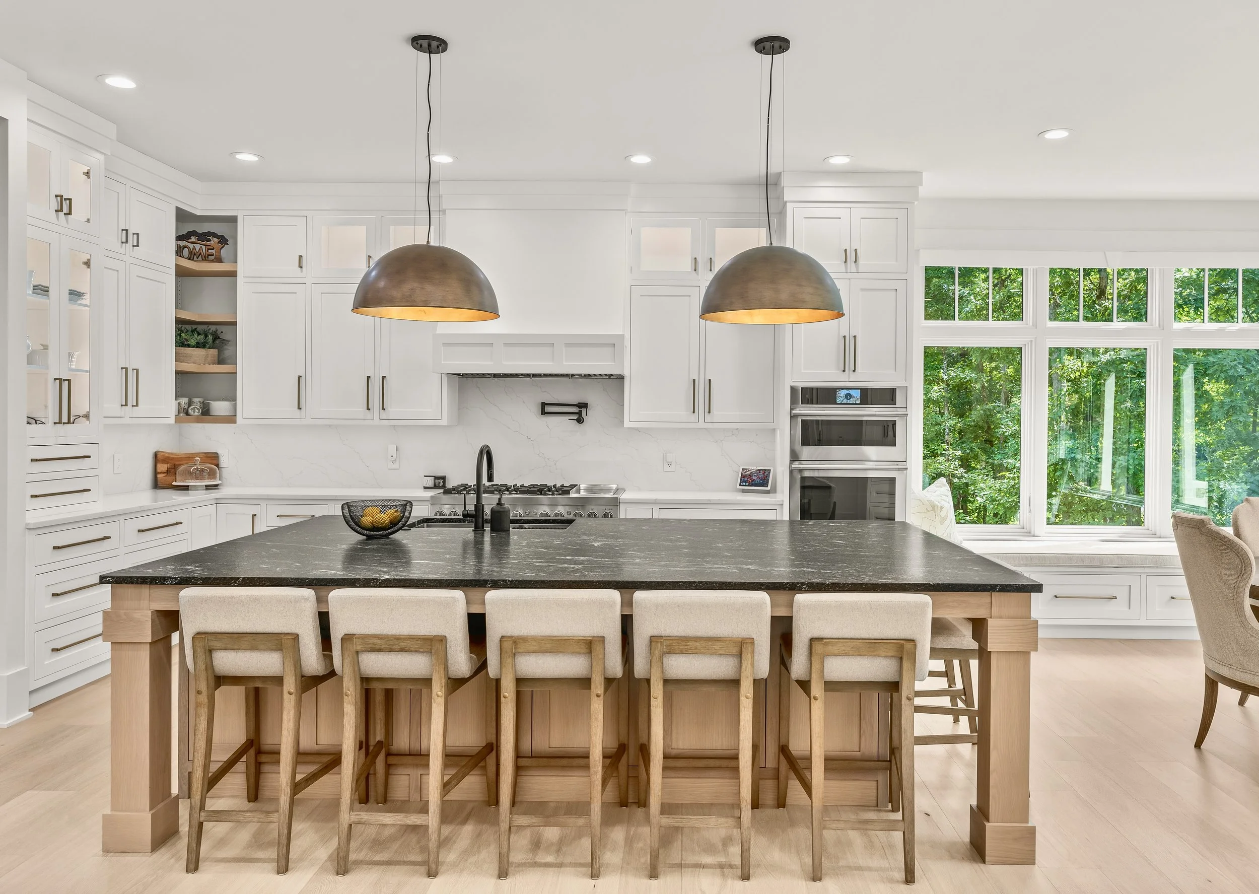 Modern white kitchen with large black countertop island, beige chairs, pendant lights, and large windows with green foliage outside.
