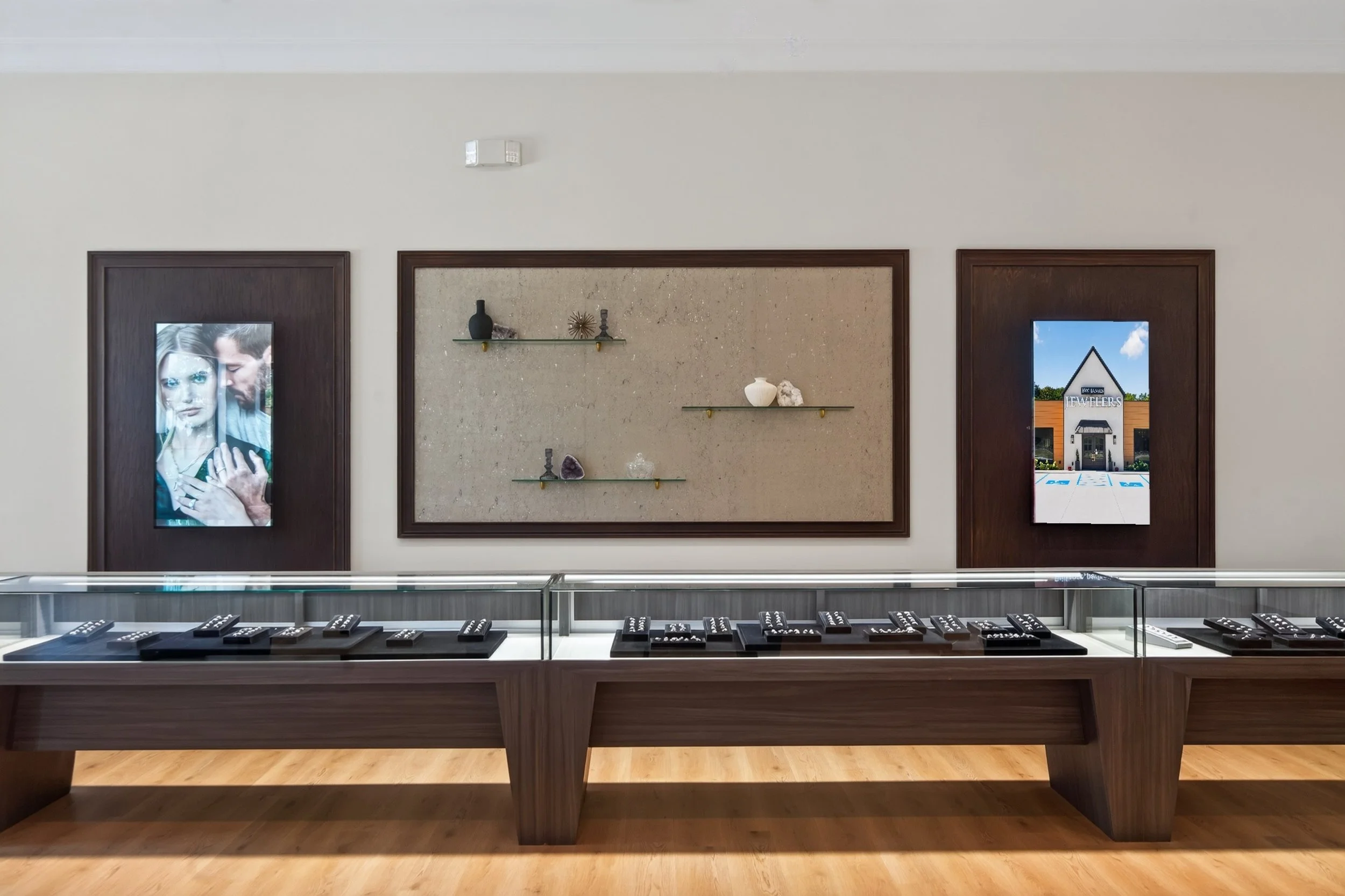 Custom jewelry display area in Cleveland, TN, featuring glass showcases, rich wood finishes, integrated digital displays, and a travertine accent wall with floating glass shelves, designed and built by local commercial builder VMH Construction.