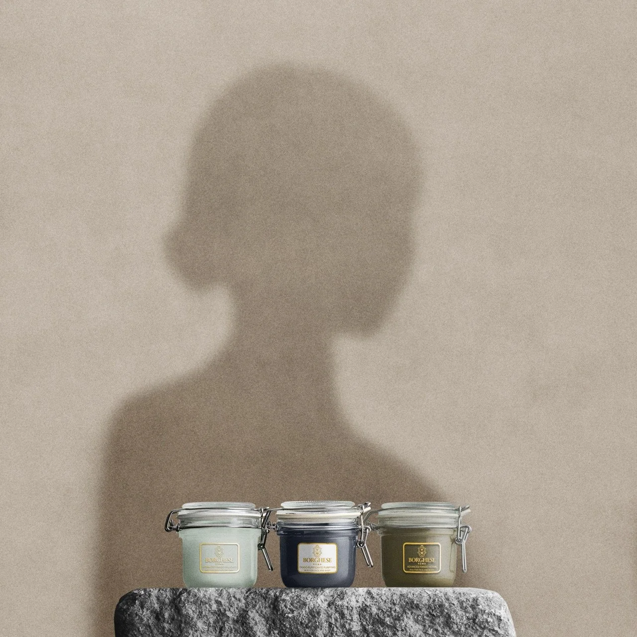Three candles in glass jars, labeled 'BORGHESE,' are placed on a textured gray stone surface. Behind them, a painted silhouette of a woman with a bun hairstyle is visible on a plain beige wall.