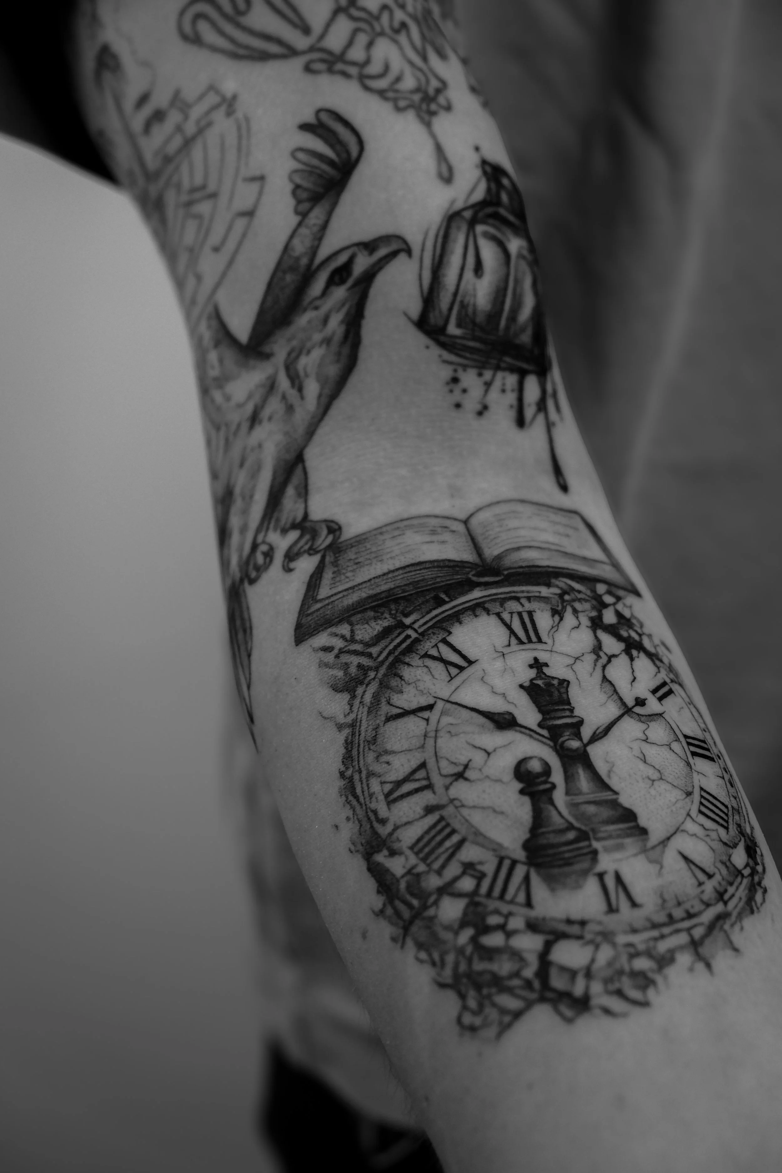 Schwarze Tätowierung auf dem Arm, zeigt eine Uhr mit römischen Zahlen, einem offenen Buch, eine Krähe und eine brennende Kerze.