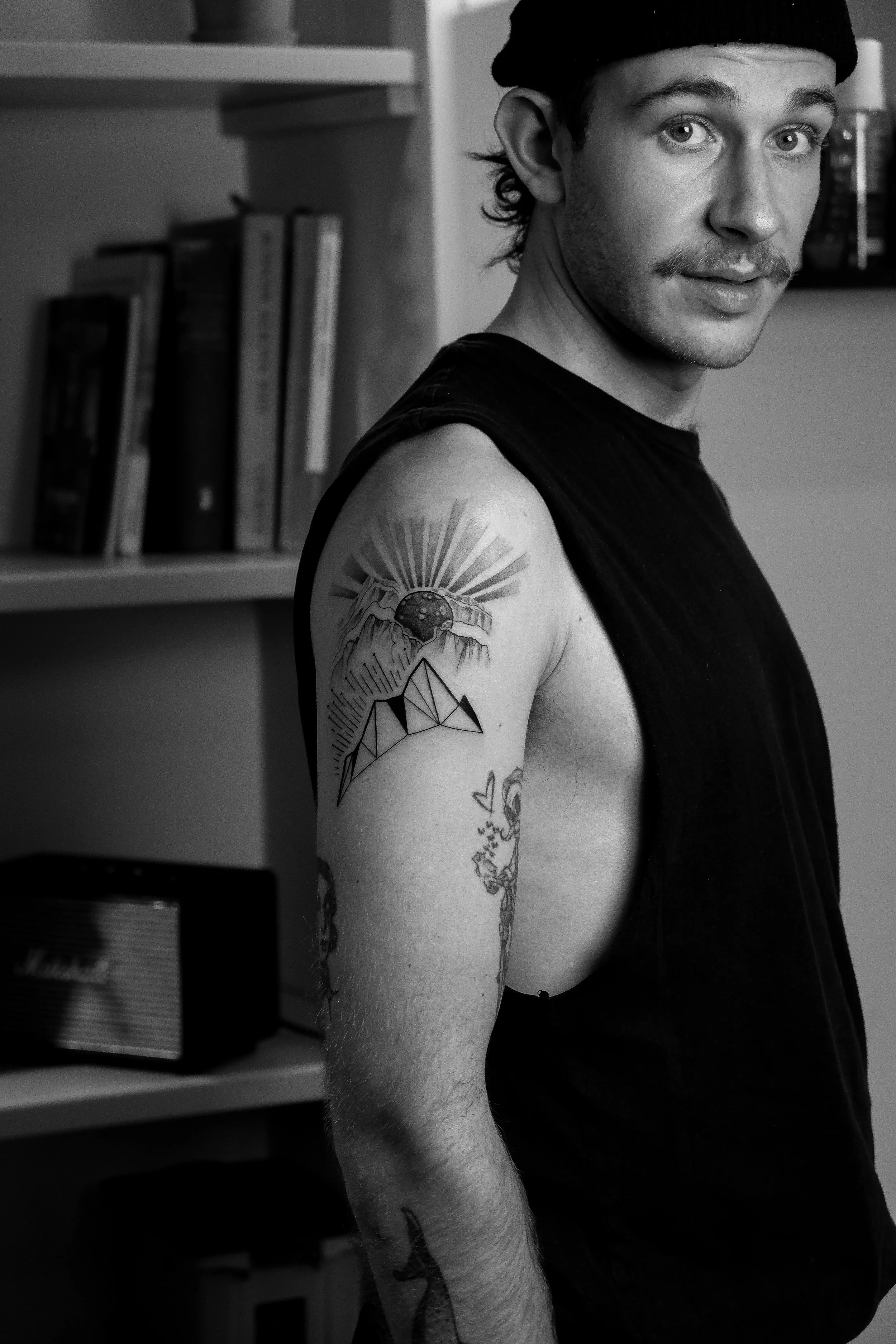 Ein junger Mann mit dunklem Haar und dunklem Oberteil zeigt sein oberarm-Tattoo in einem Raum mit Büchern und einem Marshall-Lautsprecher.