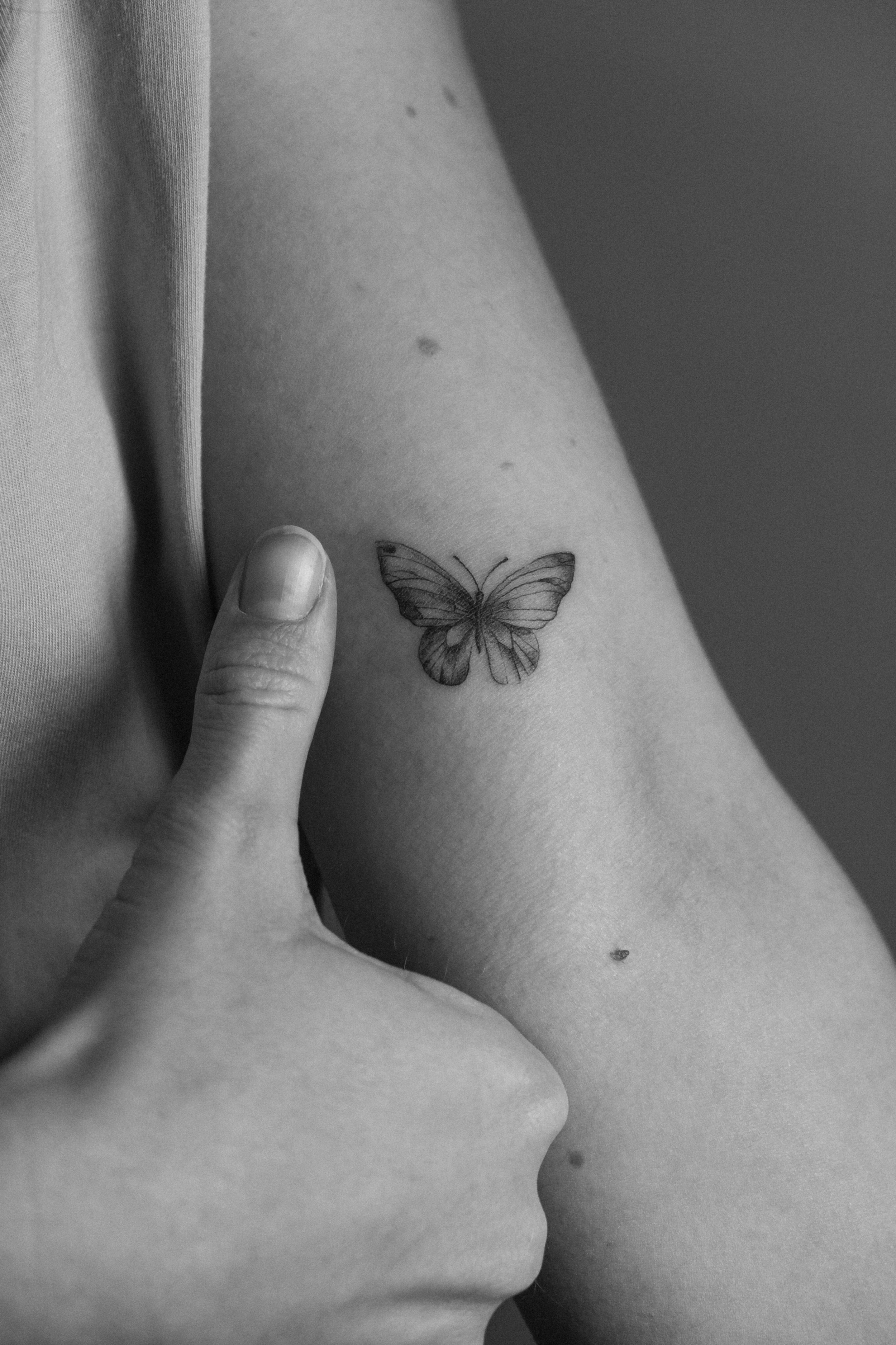Schwarz-Weiß-Foto einer Person, die eine Schmetterlings-Tätowierung am Oberarm zeigt, mit Daumen, der auf die Tätowierung zeigt.