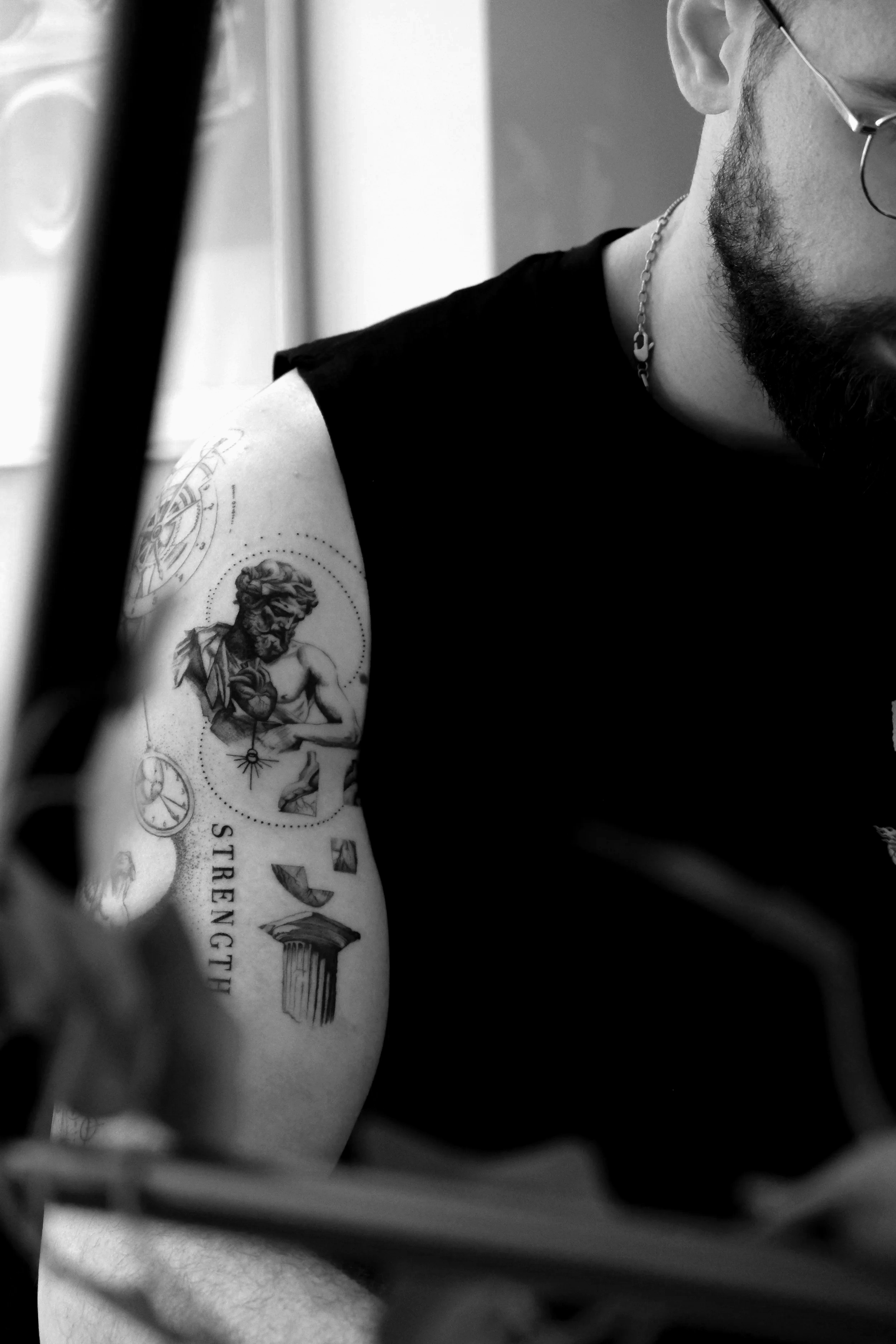 Schwarz-Weiß-Foto eines Mannes mit Tattoos am Oberarm, trägt Brille und Halskette, nur ein Teil seines Gesichts ist sichtbar.