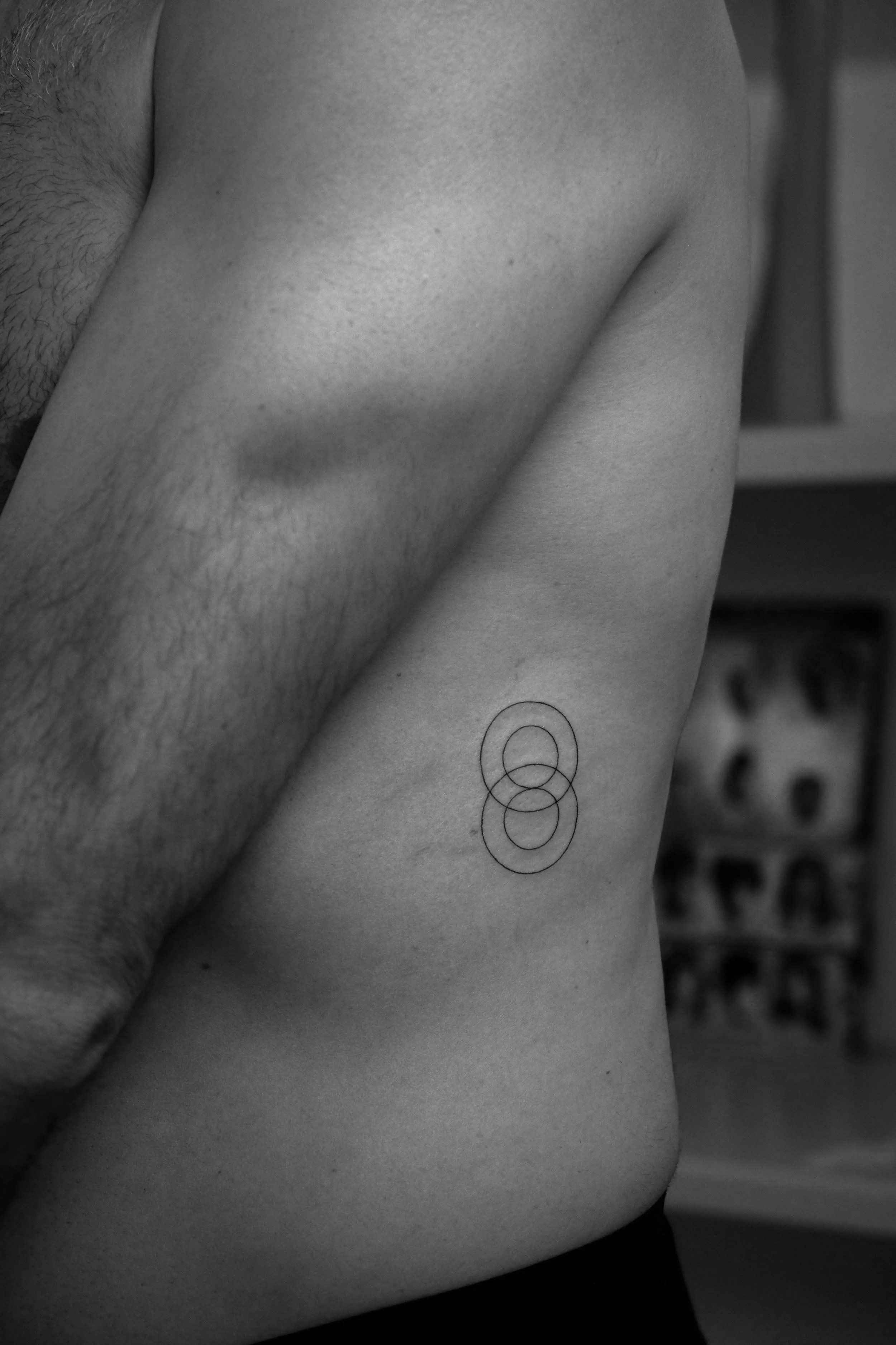 Schwarze und weiße Nahaufnahme eines männlichen Oberkörpers mit einem geometrischen Tattoo auf der Seite des Brustkorbs, im Hintergrund ein Regal mit Gegenständen.