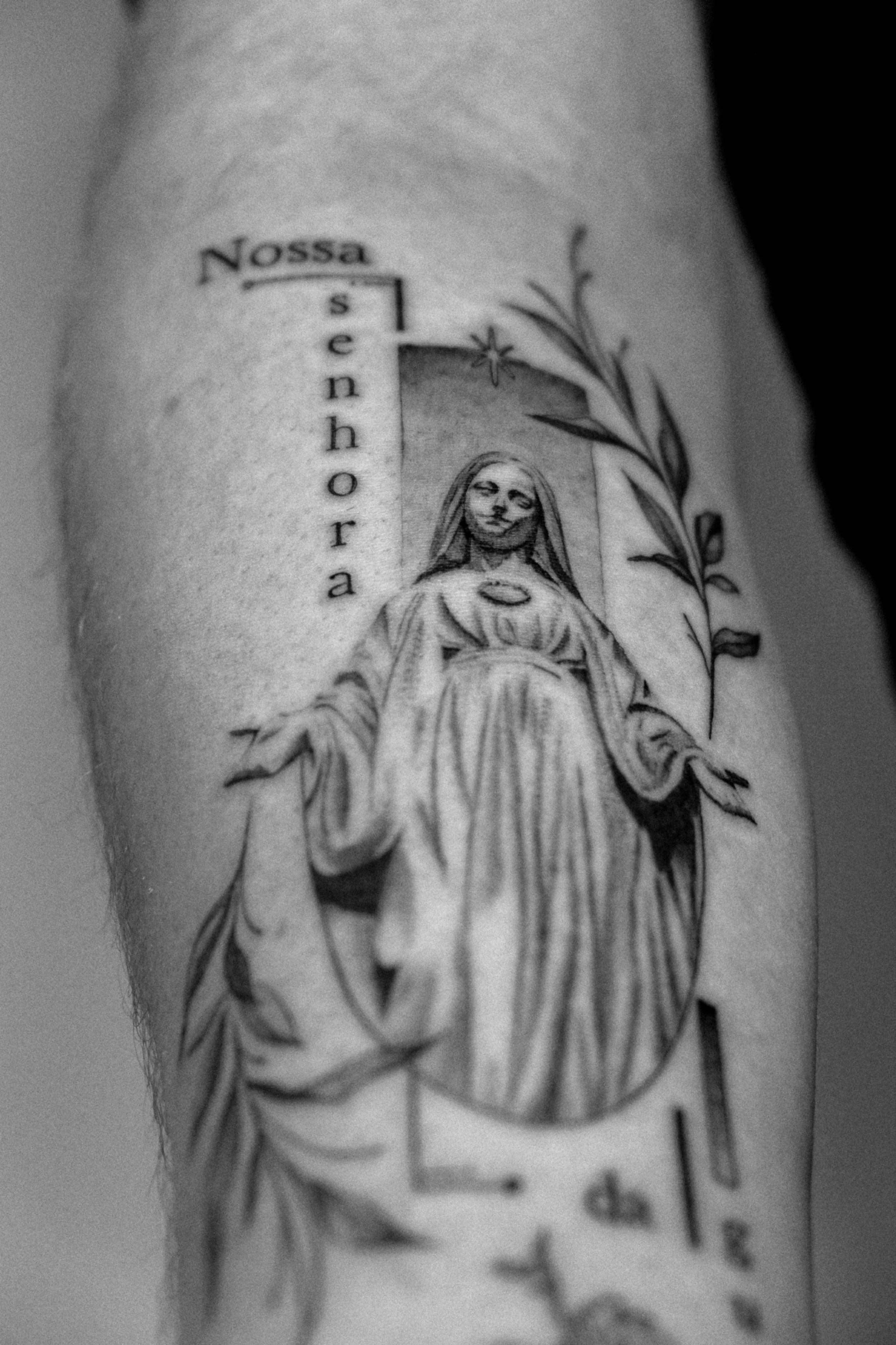 Schwarz-Weiß-Tattoo einer Heiligen mit einem Heiligenschein, umgeben von Blättern, mit dem Text "Nossa senhora".