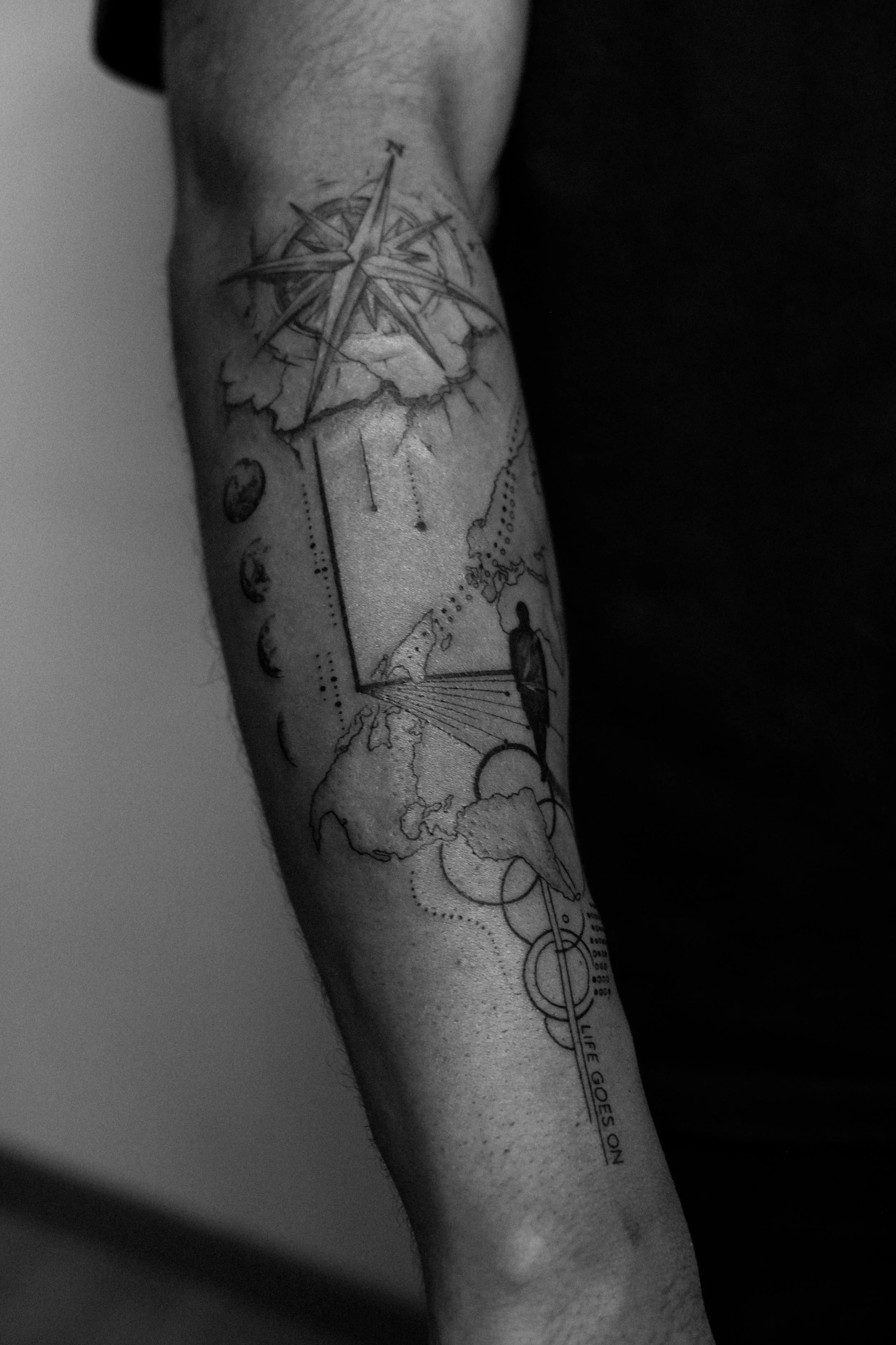 Tattoo auf einem Arm, das eine Weltkarte, einen Kompass, eine Sonne, einen Mond, einen Menschen, der in Richtung Sonne schaut, und Textenthält. 