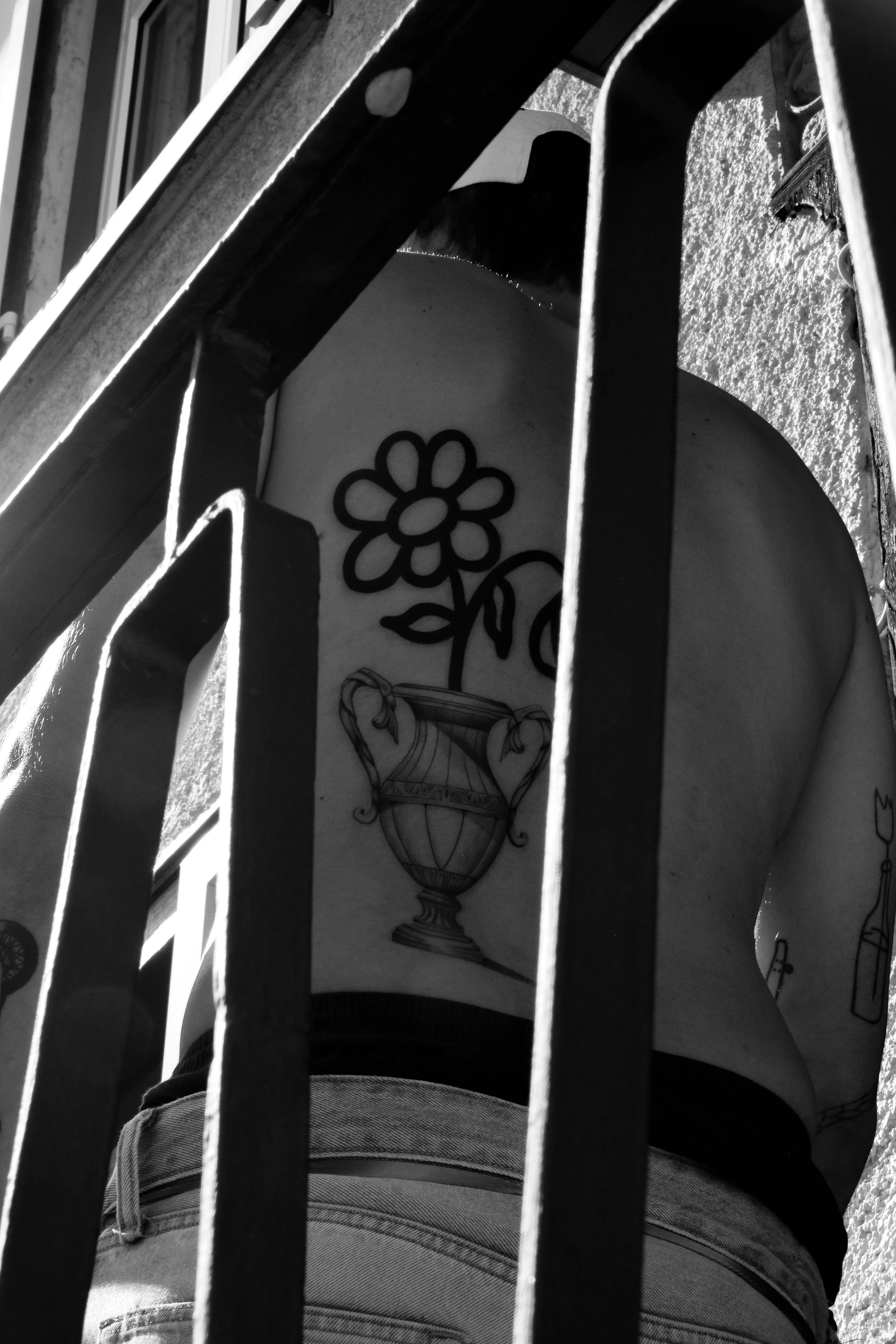 Schwarz-weiß-Foto eines Tattoos auf der Schulter einer Person, sichtbar durch Metallstangen eines Geländers, das den Blick versperrt. Das Tattoo zeigt eine Blume und einen antiken Krug.