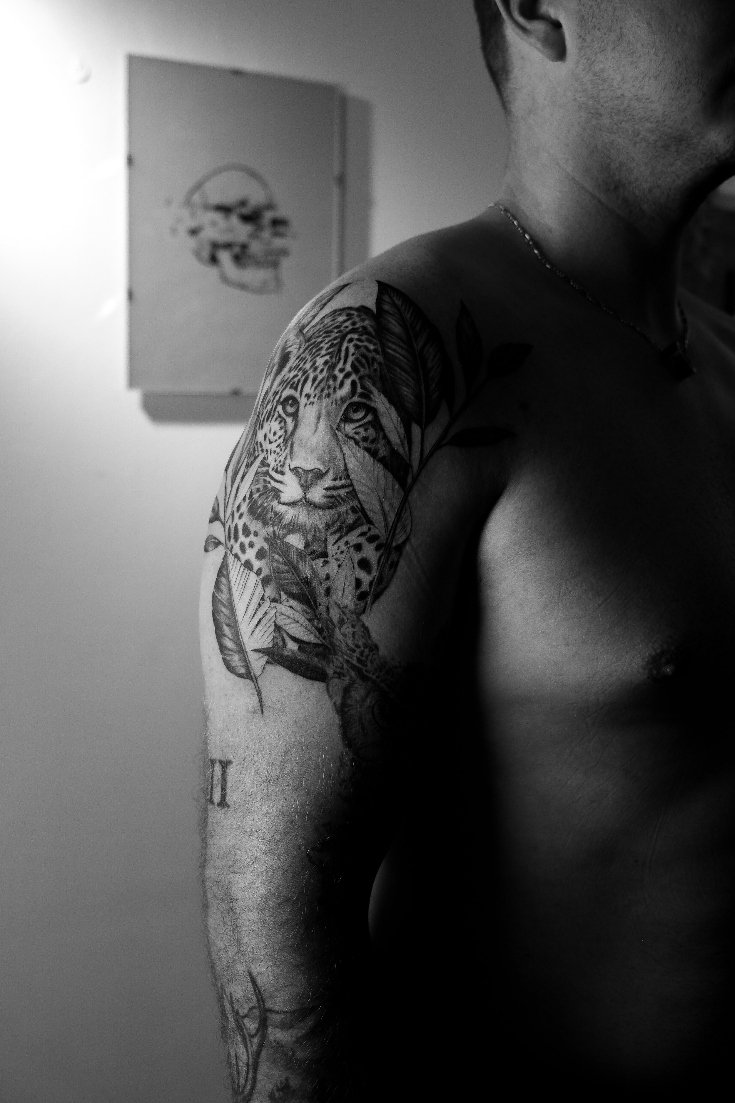 Schwarze und weiße Tattoo eines Leopardenkopfes auf der Schulter eines Mannes, mit Blättern und Tieren, im Hintergrund eine Skulptur eines menschlichen Schädels.