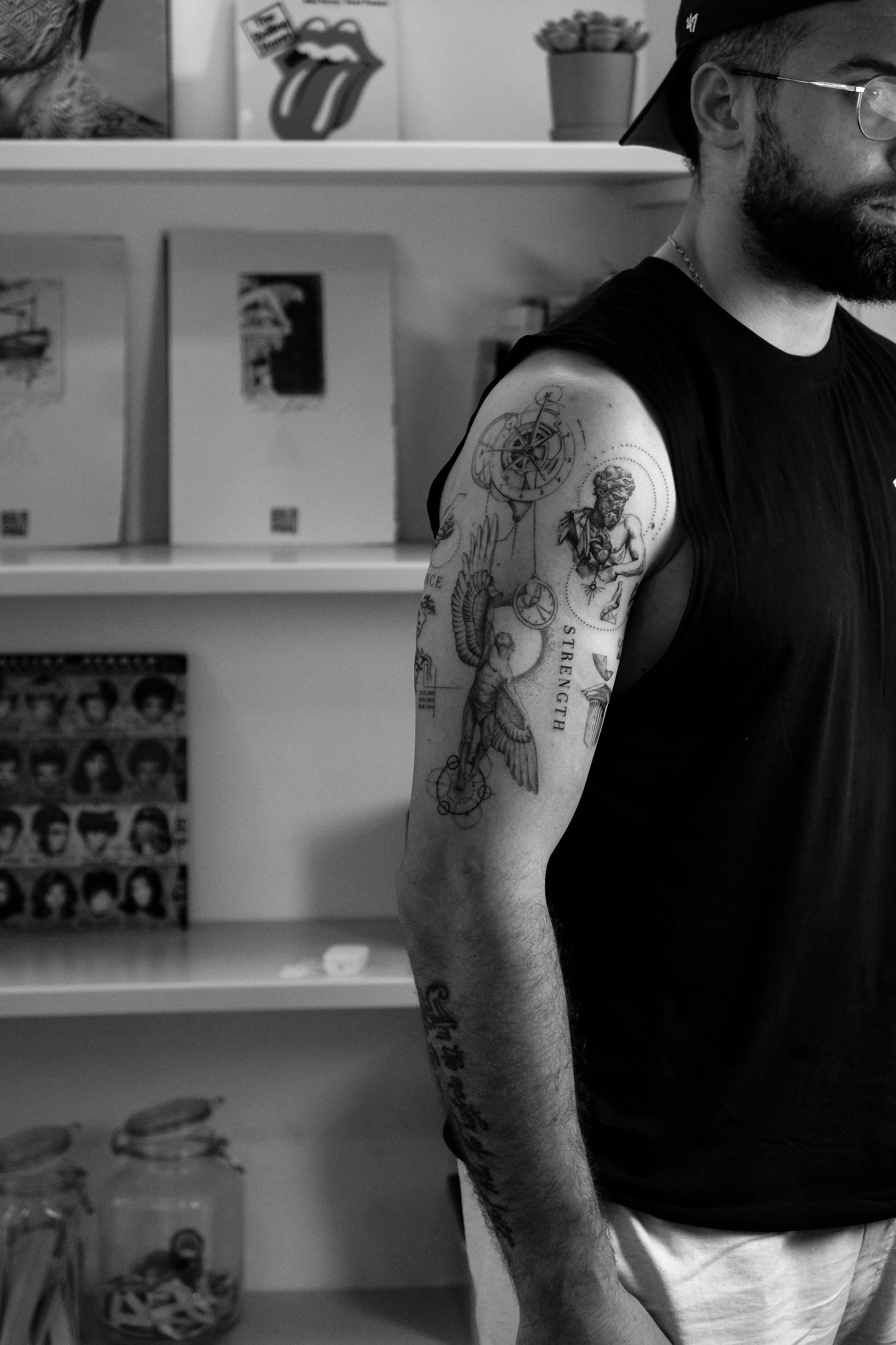 Ein Mann mit detaillierten Black & Grey Tattoos auf dem Arm, man sieht nur den Teil seines Gesichts, trägt eine schwarze Weste und eine schwarze Mütze. Im Hintergrund sind Bücher, Poster und Dekorationsgegenstände sichtbar.