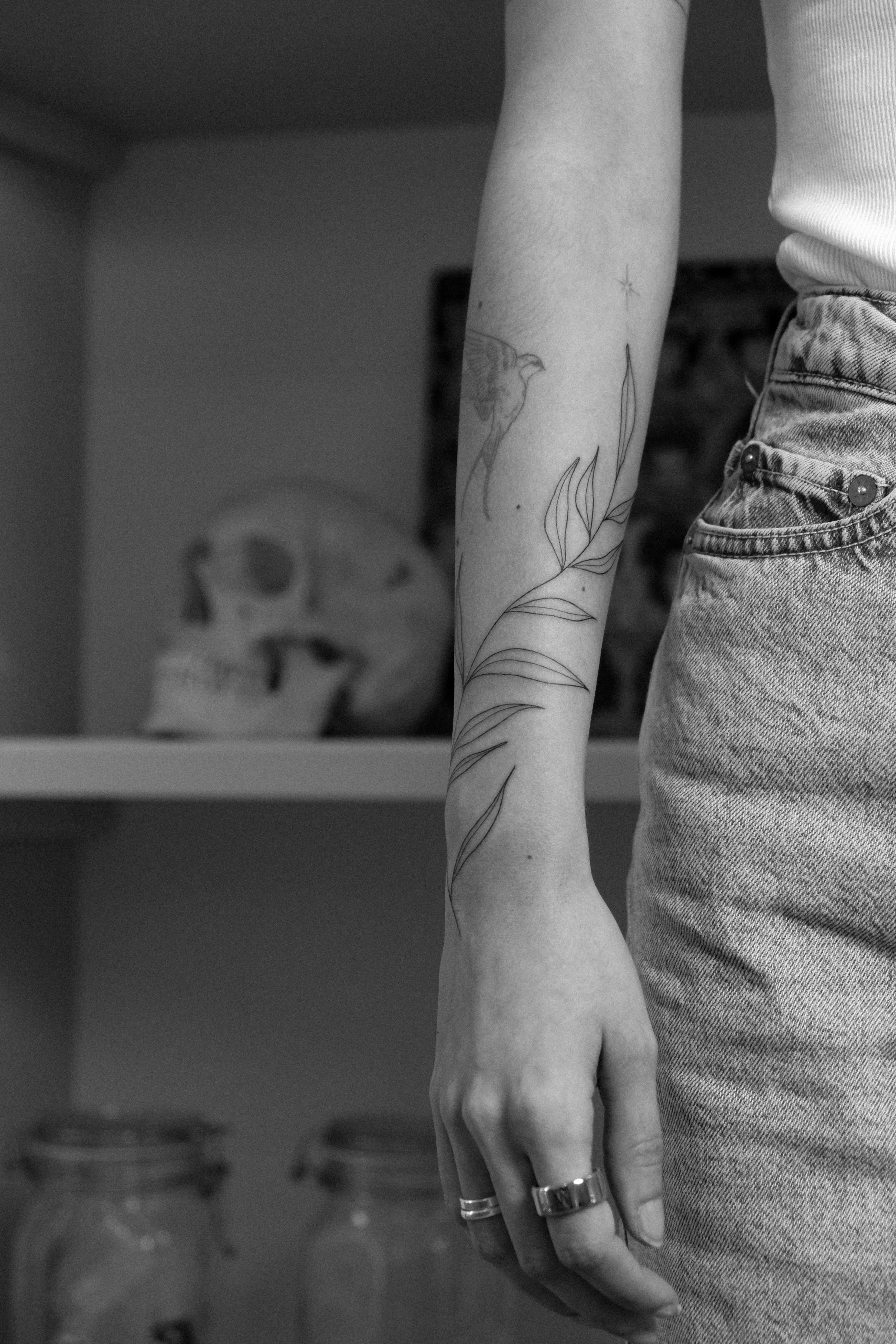 Ein Arm mit Tattoo, der eine Pflanze und einen Vogel zeigt, trägt mehrere Ringe. Im Hintergrund ist ein Puzzlekopf auf einem Regal zu sehen.