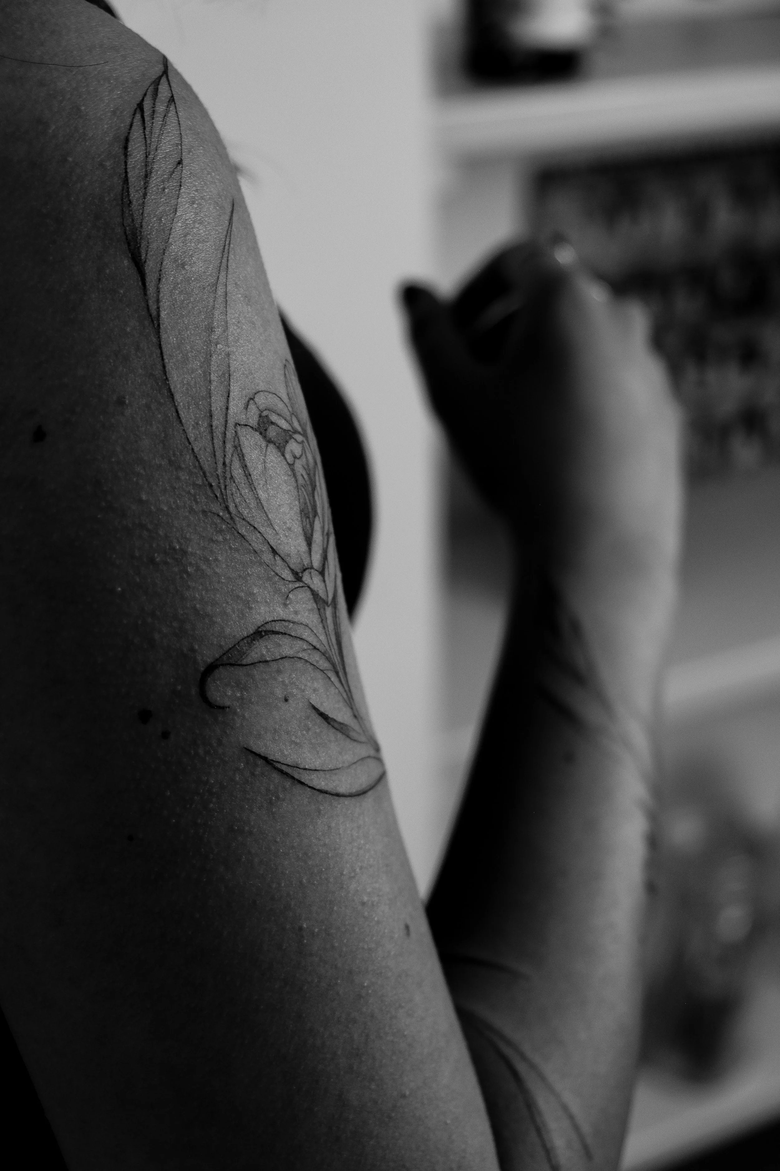 Schwarz-weiß Foto eines Arms mit Tattoo, das auf einen Stift oder Marker hält, vermutlich beim Tätowieren im Inneren einer Raum mit Schränken im Hintergrund.