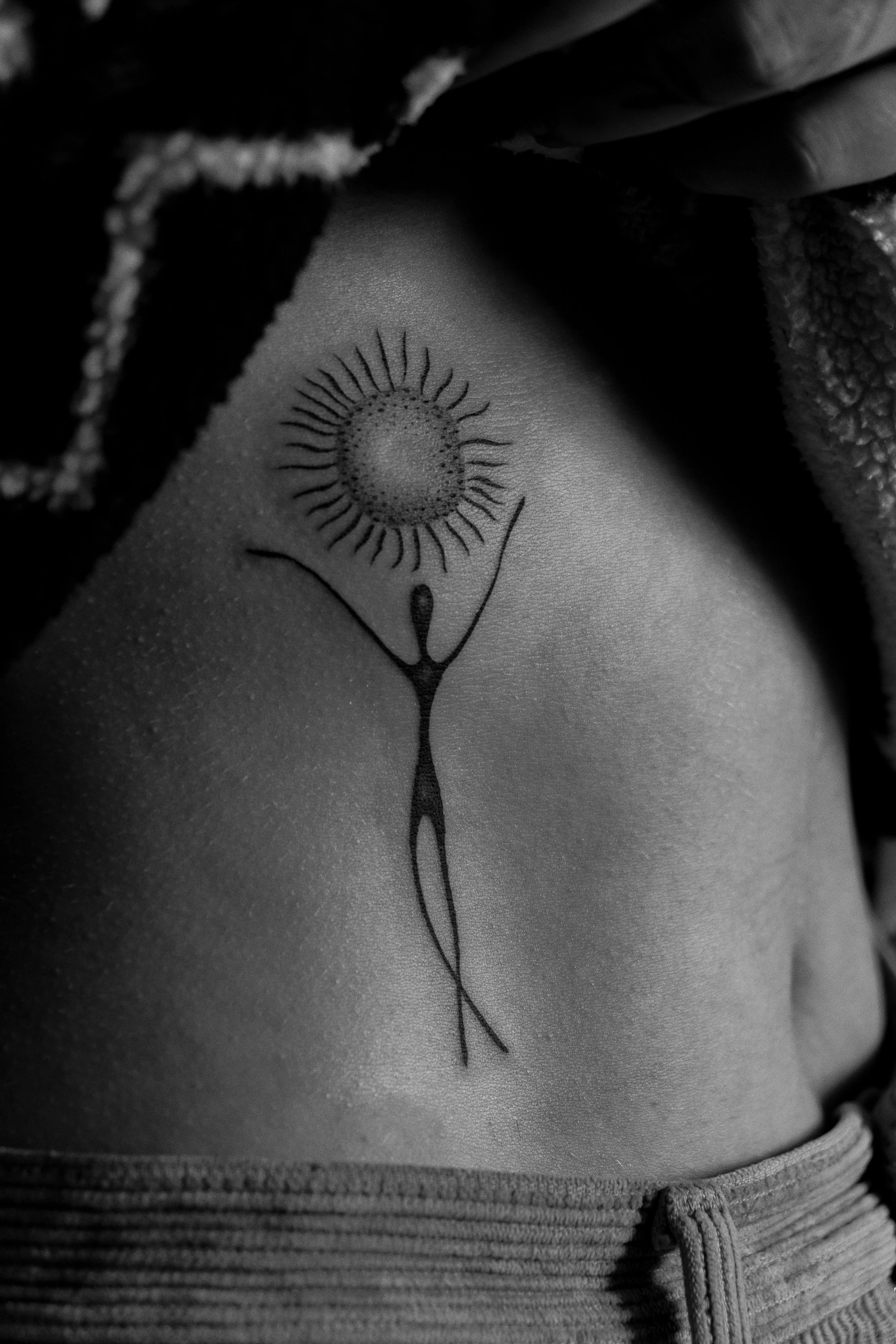 Schwarz-weiß-Tattoo eines stilisierten Menschen mit erhobenen Armen, das eine Sonne oder Blume hält, auf einem Arm. Die Person ist auf der Seite liegend und trägt eine Hose.